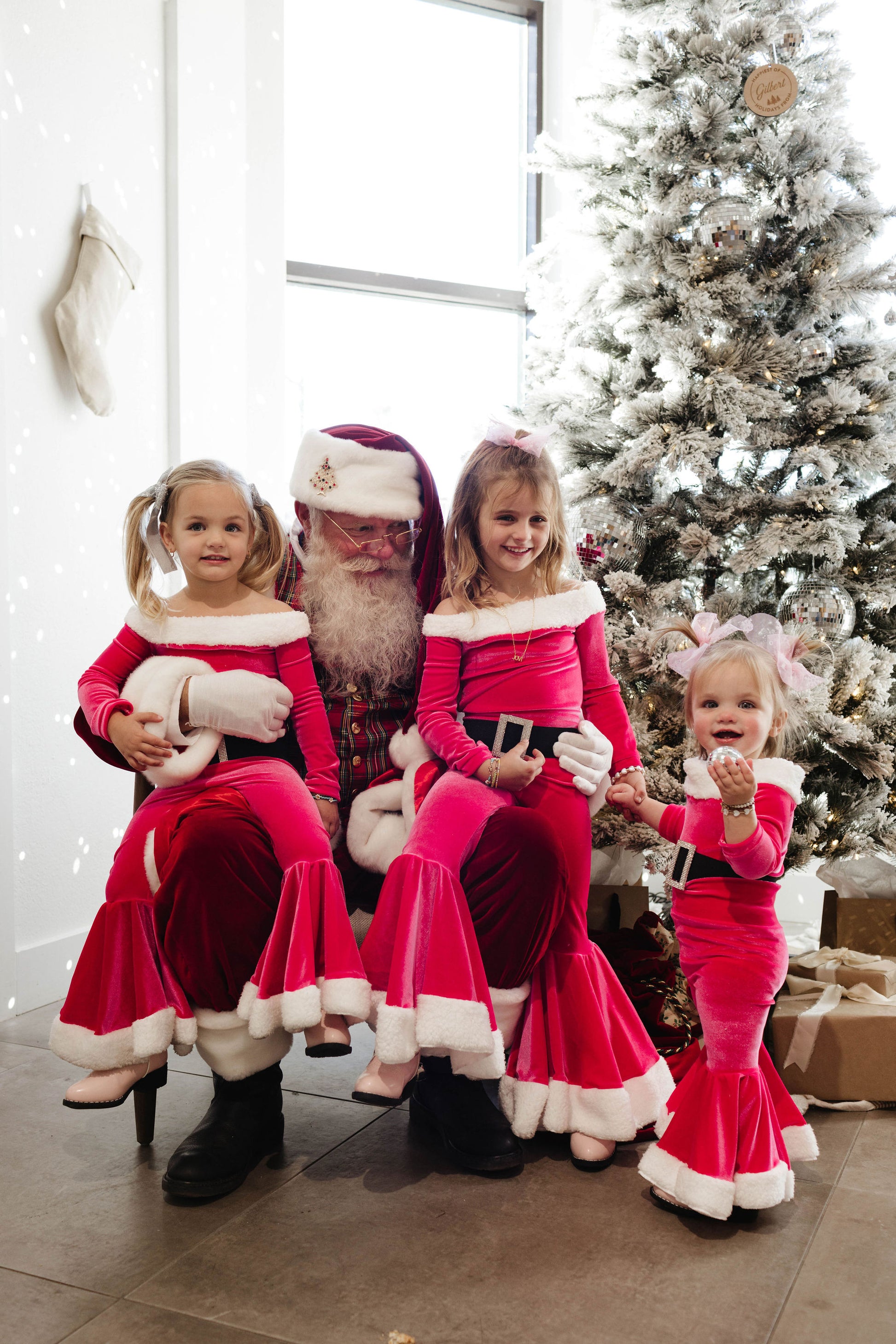 Three children, dressed in matching red and white outfits from Forever French Baby, pose with Santa Claus in front of a decorated Christmas tree. The children are smiling and holding Santa's arms while he is seated. This festive scene, beautifully captured by Amanda Riley Photos, showcases the holiday spirit with a room filled with decorations and presents under the tree. This charming moment is part of the Santa Photos | Forever French Baby x Amanda Riley Photos collection by Amanda Riley Photos.