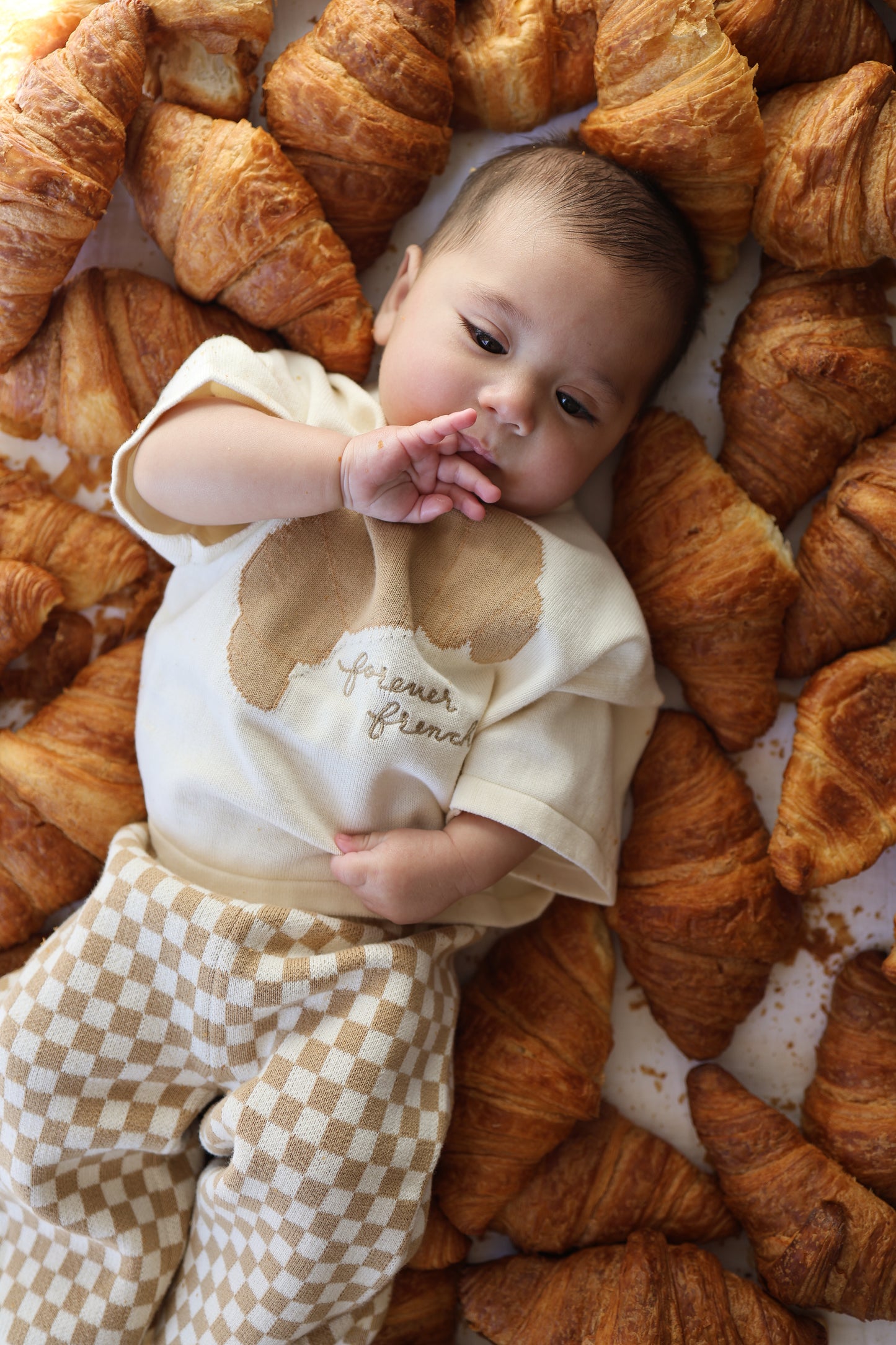 A baby dressed in the forever french baby Knit Pant Set | Croissants lies among croissants, touching their face and gazing upward.