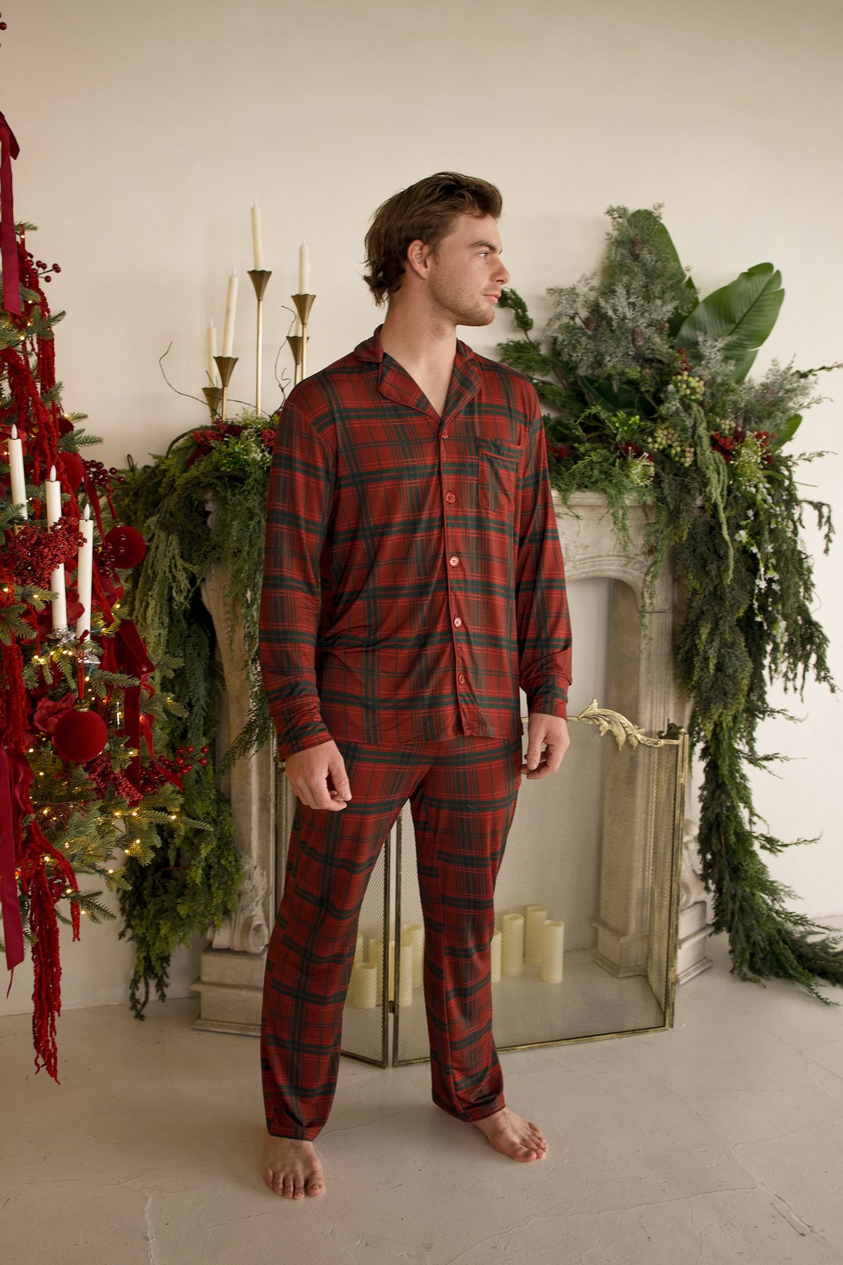 A man wearing a Men’s Bamboo Button Up Lounge Set | Christmas Plaid in red and green, standing in front of a fireplace with holiday decorations.