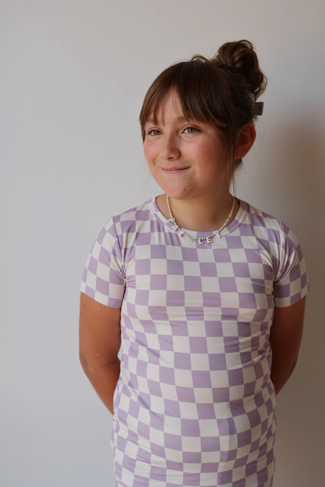 A young girl with brown hair in a bun smiles softly, wearing the forever french baby Bamboo Short Set in Grape Soda and a beaded necklace, standing against a plain light background.