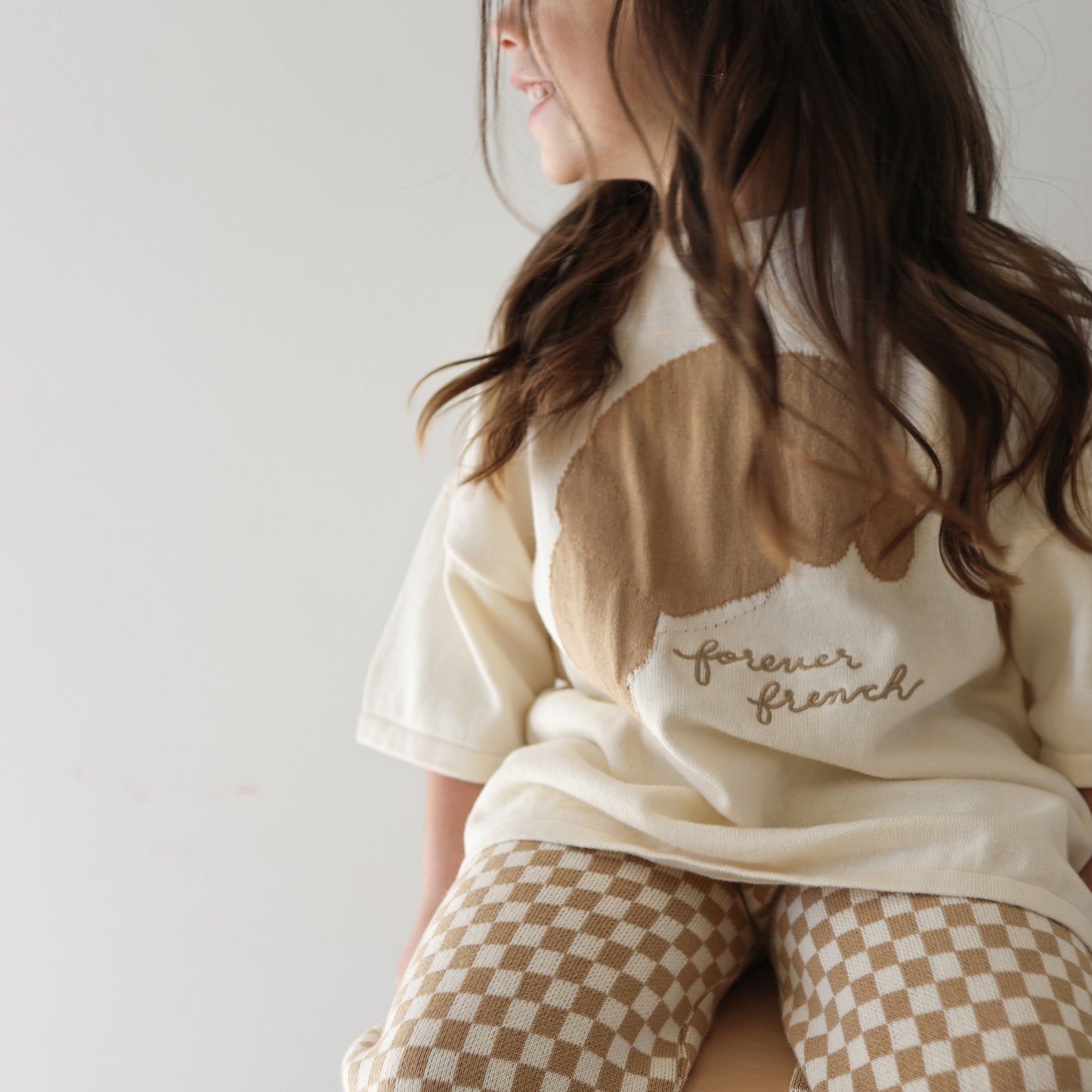 A young girl with long brown hair smiles while sitting on a stool, wearing the Knit Pant Set | Croissants from forever french baby, featuring an abstract top and brown and white checkered pants.