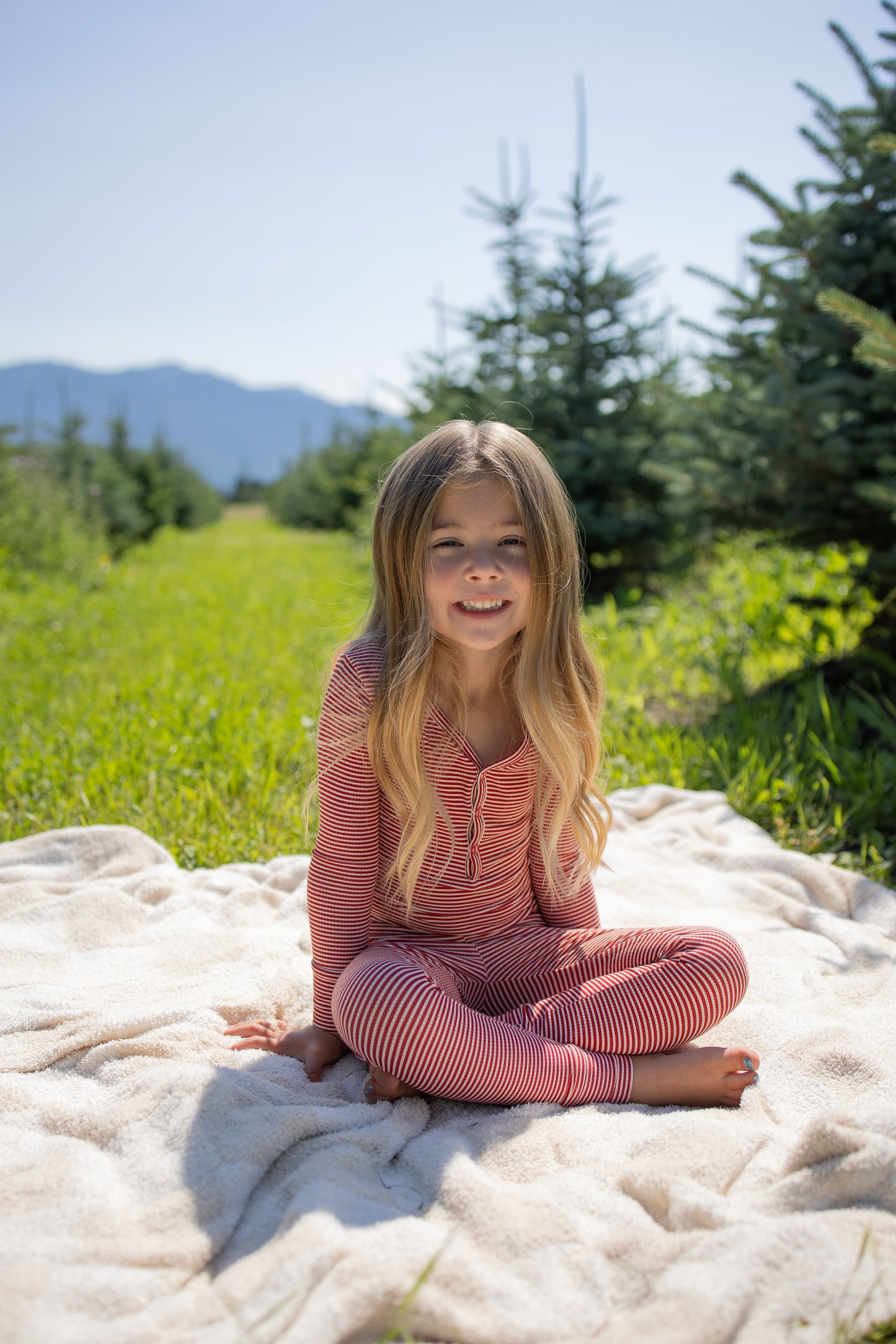 A young child with long hair sits barefoot on a blanket outdoors, wearing the forever french baby Bamboo Two Piece Pajamas in Peppermint Stripe Ribbed, surrounded by green grass and pine trees, with mountains visible in the background under a clear sky.
