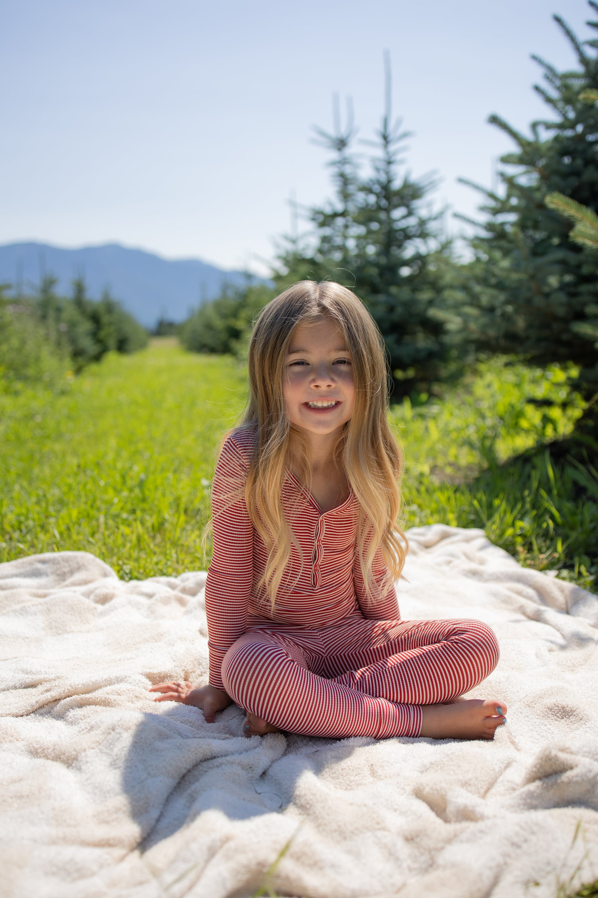 A young child with long hair sits barefoot on a blanket outdoors, wearing the forever french baby Bamboo Two Piece Pajamas in Peppermint Stripe Ribbed, surrounded by green grass and pine trees, with mountains visible in the background under a clear sky.