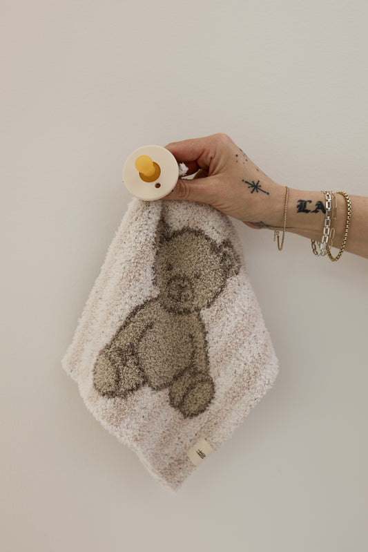 A tattooed hand with bracelets holds a forever french baby Lovey | Teddy—a pacifier attached to a plush microfiber lovey with an embroidered teddy bear—against a plain, light-colored wall.