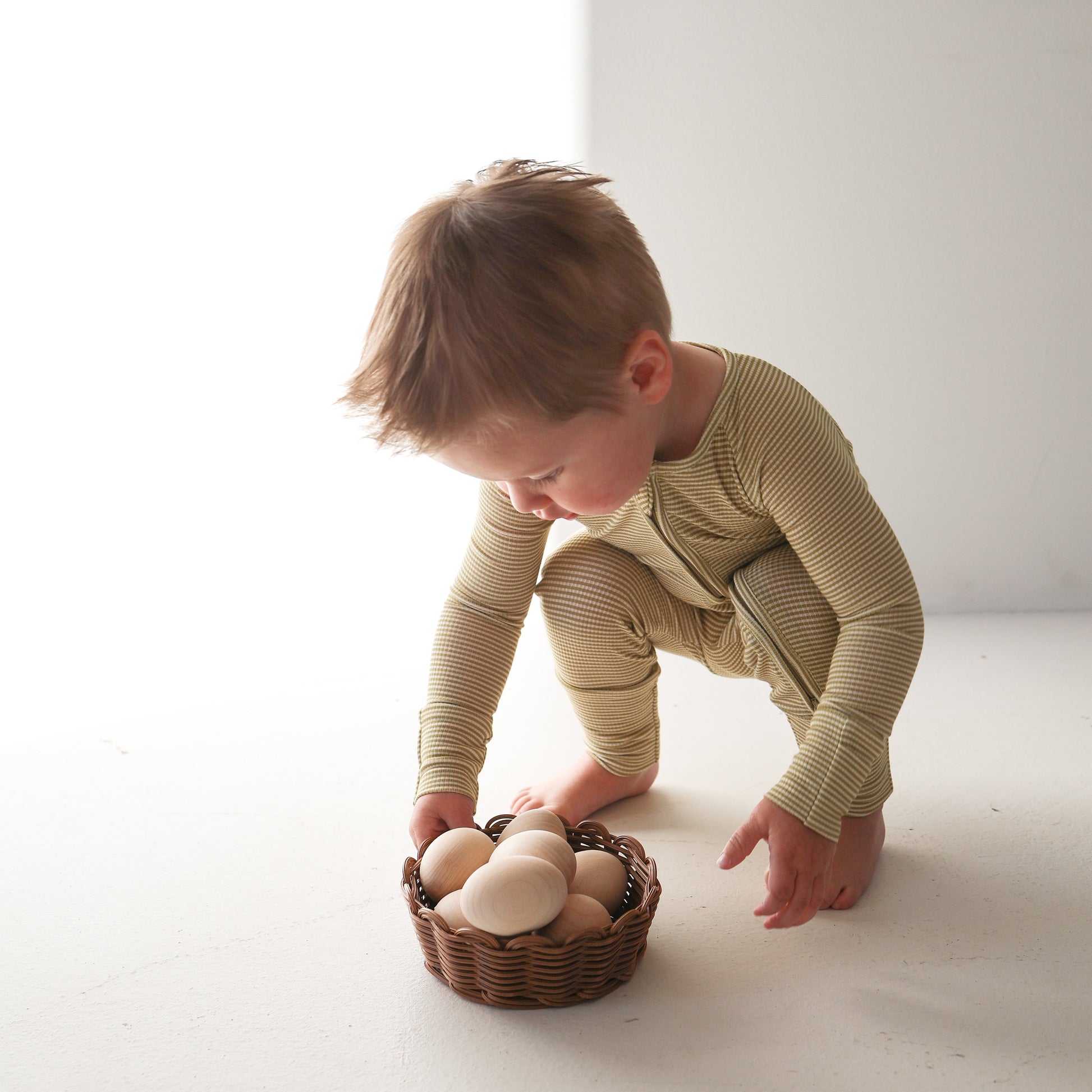 A young child wearing forever french baby’s Willow Stripe Ribbed Bamboo Zip Pajamas squats on a white floor, reaching for a wicker basket with wooden eggs in soft, natural light.