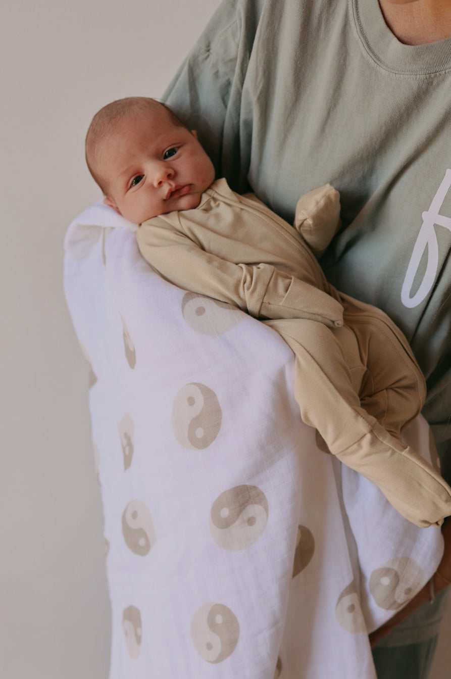 A baby dressed in forever french baby's Bamboo Zip Pajamas in Limestone rests in the arms of an adult wearing a green shirt. The baby is wrapped in hypo-allergenic clothing, specifically a white blanket adorned with beige yin-yang symbols.