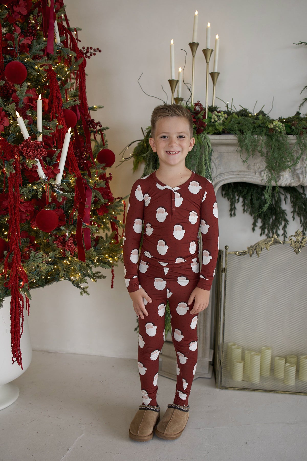 A child stands in front of a Christmas tree wearing matching Bamboo Two Piece Pajamas | Vintage Santa, featuring a burgundy long-sleeve design with a repeating white Santa Claus print.