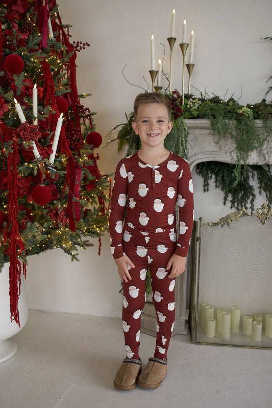 A child stands in front of a Christmas tree wearing matching Bamboo Two Piece Pajamas | Vintage Santa, featuring a burgundy long-sleeve design with a repeating white Santa Claus print.