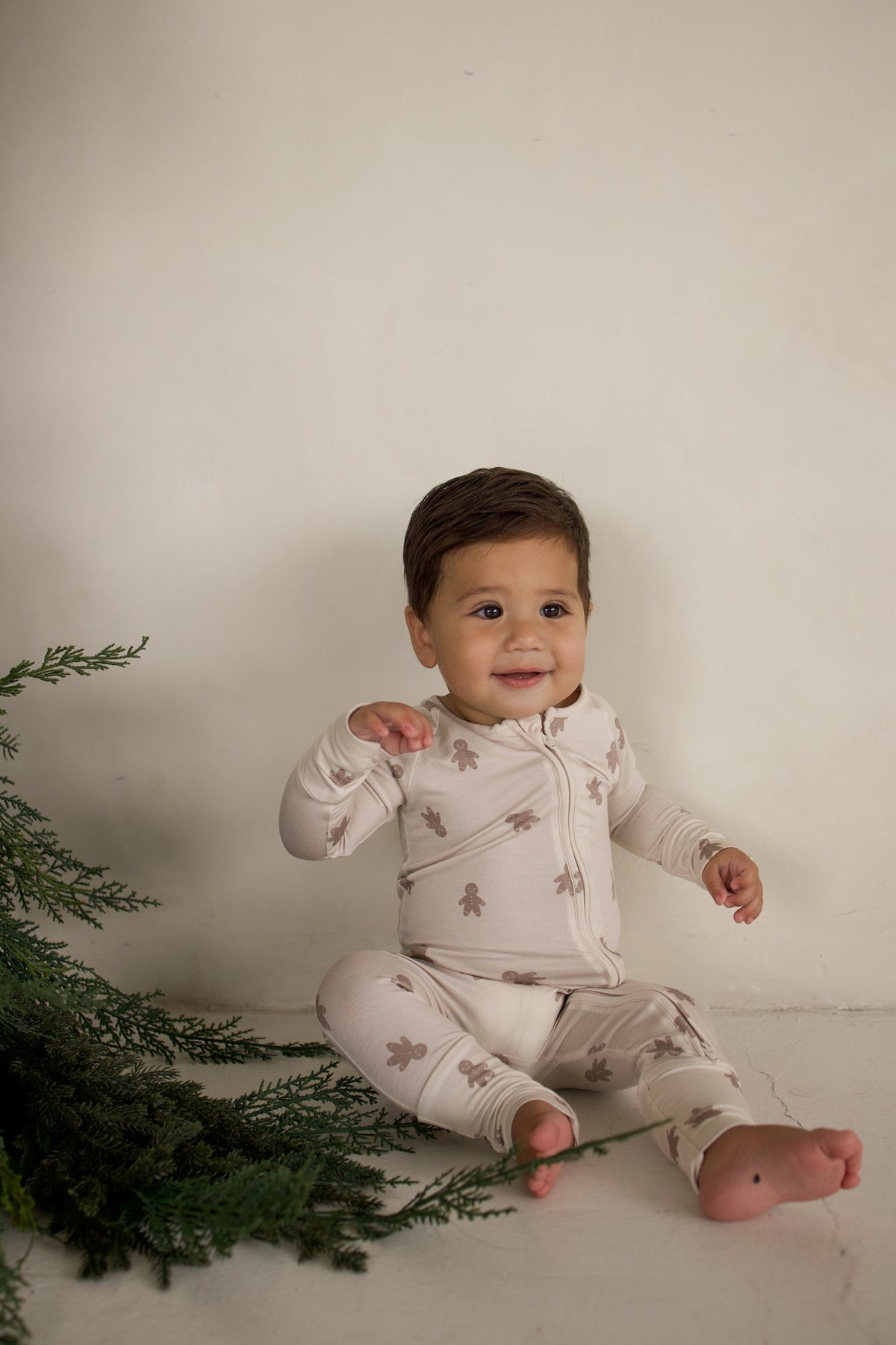 A baby sits on the floor wearing Bamboo Zip Pajamas | Gingersnap patterned with small gingerbread men.