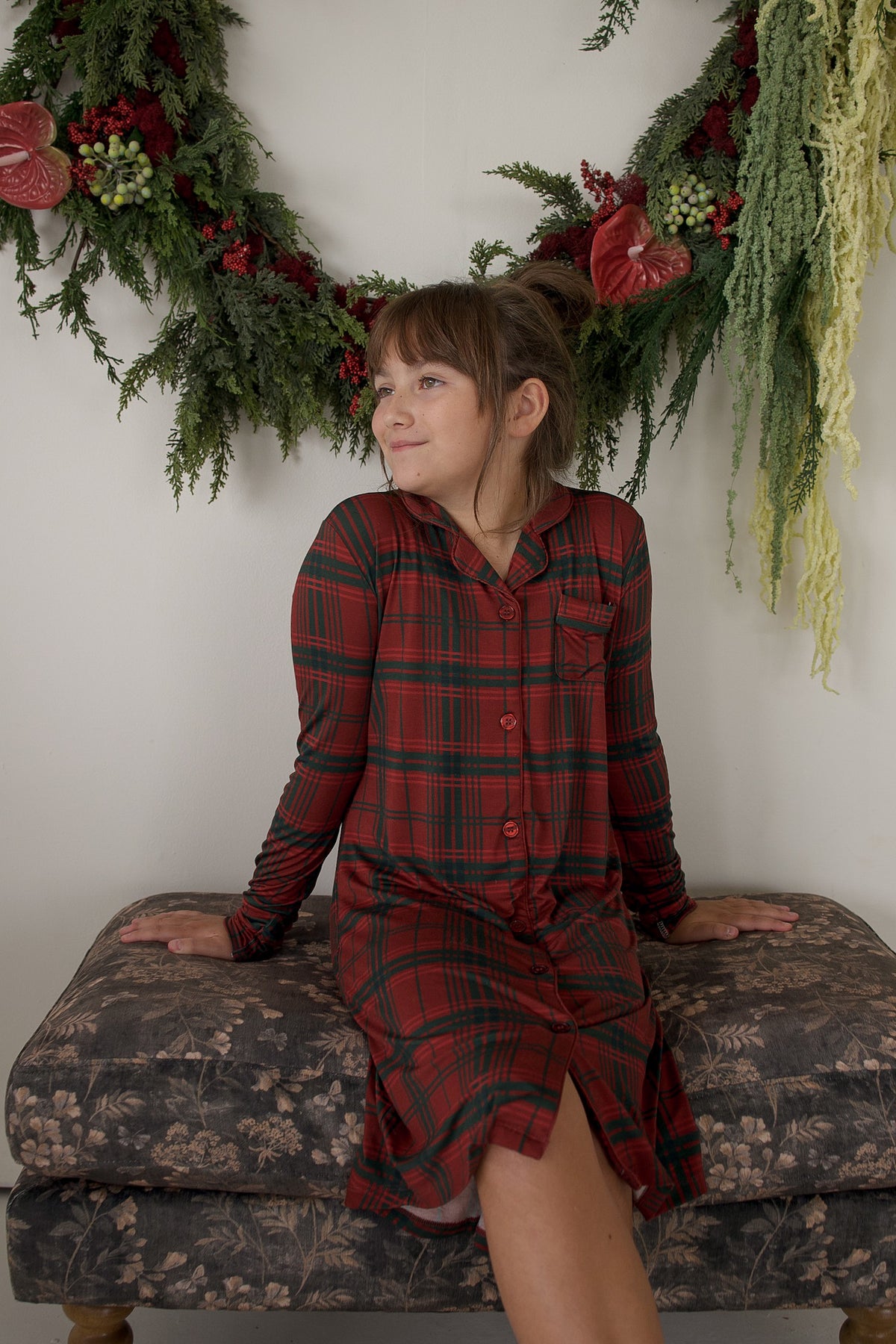 A young girl wears the Bamboo Sleeping Dress | Christmas Plaid by forever french baby, seated on a floral bench beside a green and red holiday wreath hanging on a white wall.