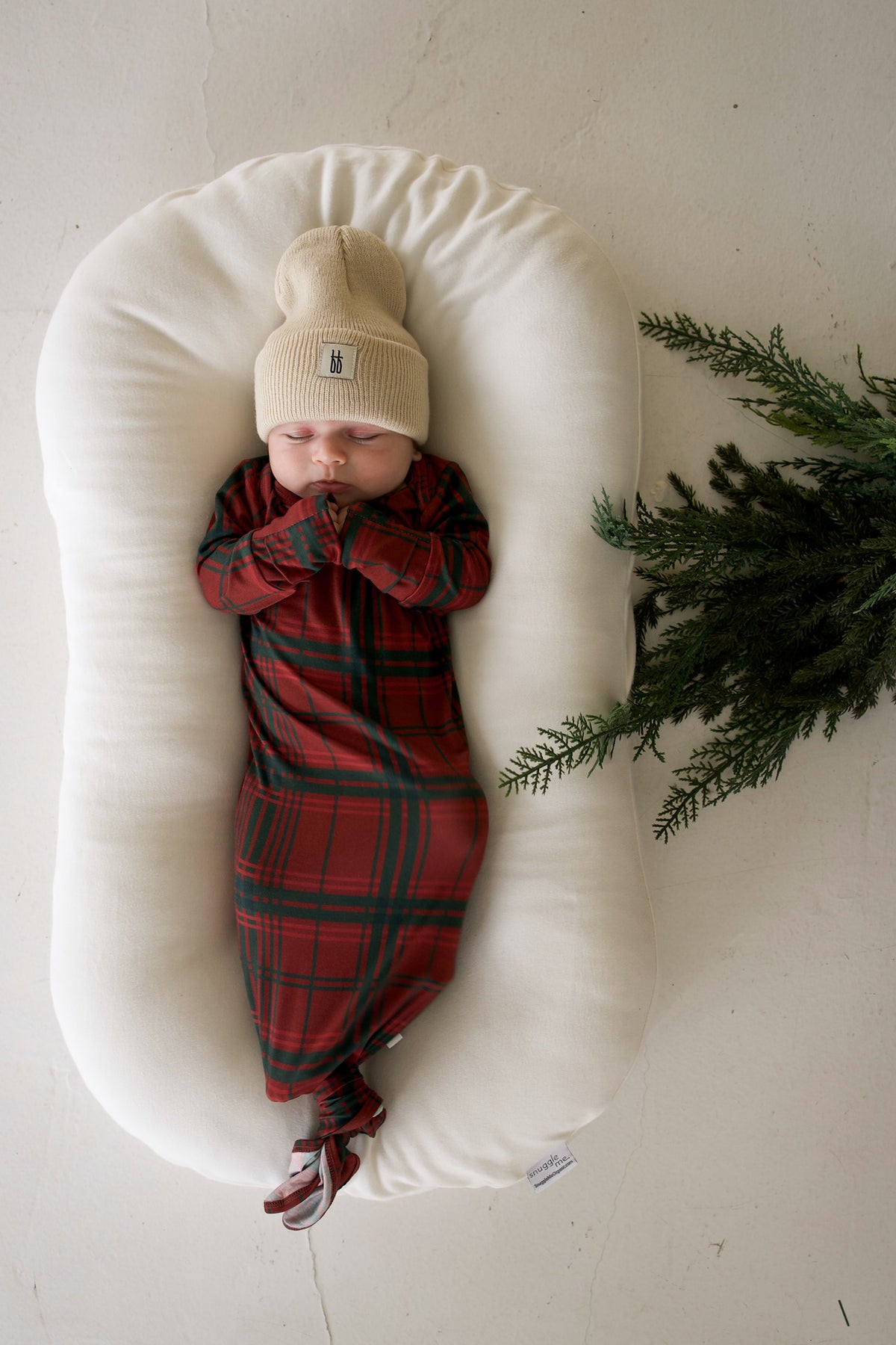 A sleeping baby in a Bamboo Knotted Gown | Christmas Plaid and a beige beanie, resting in a cream-colored infant lounger next to a pine branch.