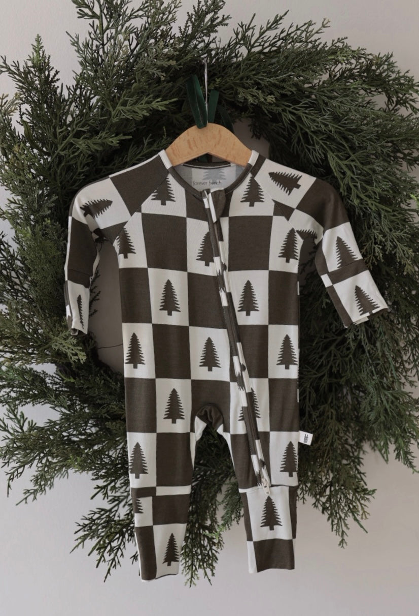 The forever french baby Bamboo Zip Pajamas in Christmas Tree Checkerboard—hypoallergenic and breathable with a brown and white checkered pine tree print—hang on a wooden hanger in a green pine wreath, perfect cozy sleepwear for your baby.