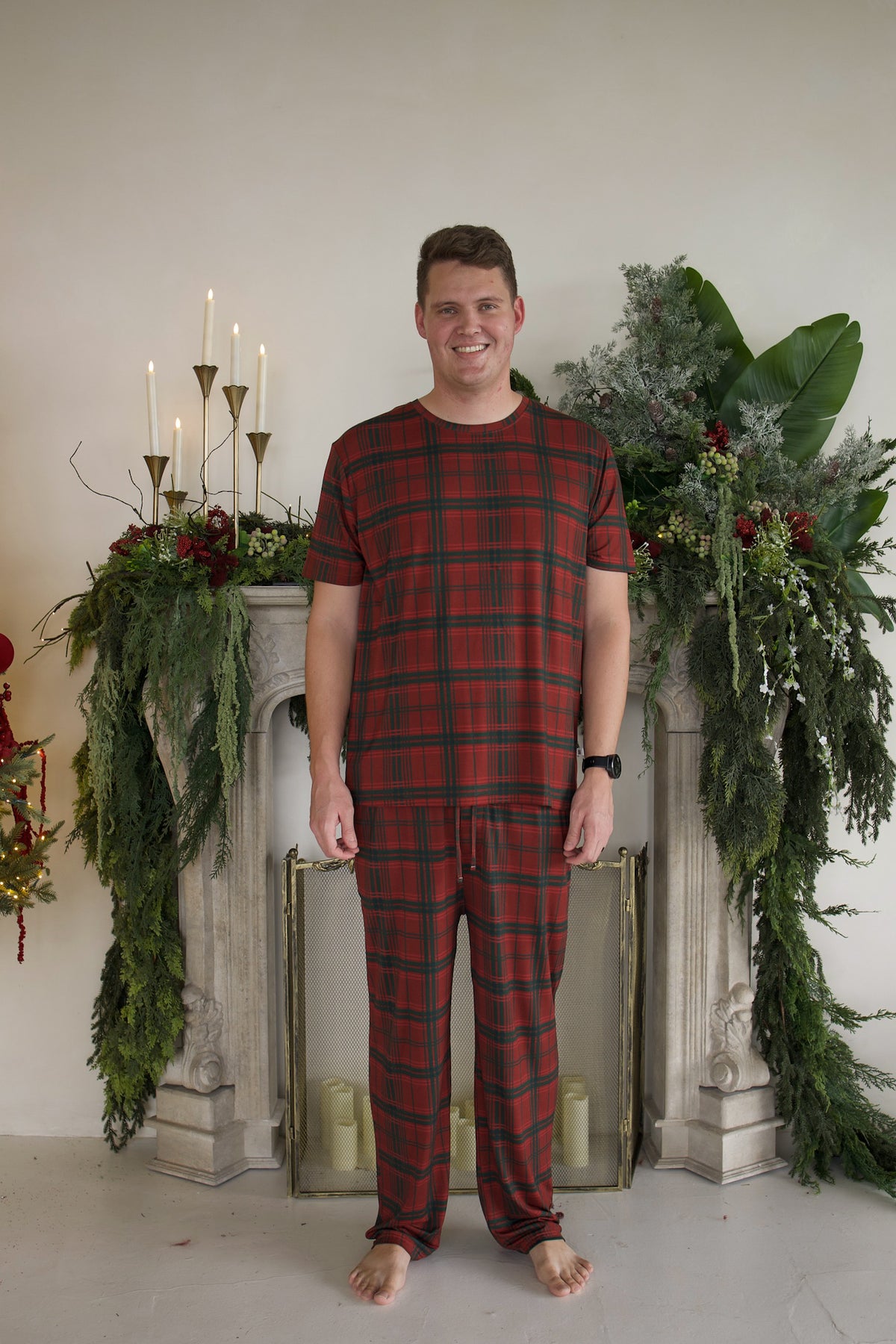 A smiling man wears a red and green Men’s Short Sleeve Pajamas | Christmas Plaid set while standing in front of a fireplace decorated with greenery and candles.