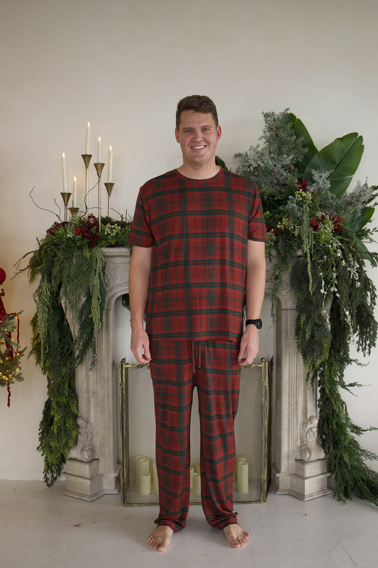 A smiling man wears a red and green Men’s Short Sleeve Pajamas | Christmas Plaid set while standing in front of a fireplace decorated with greenery and candles.