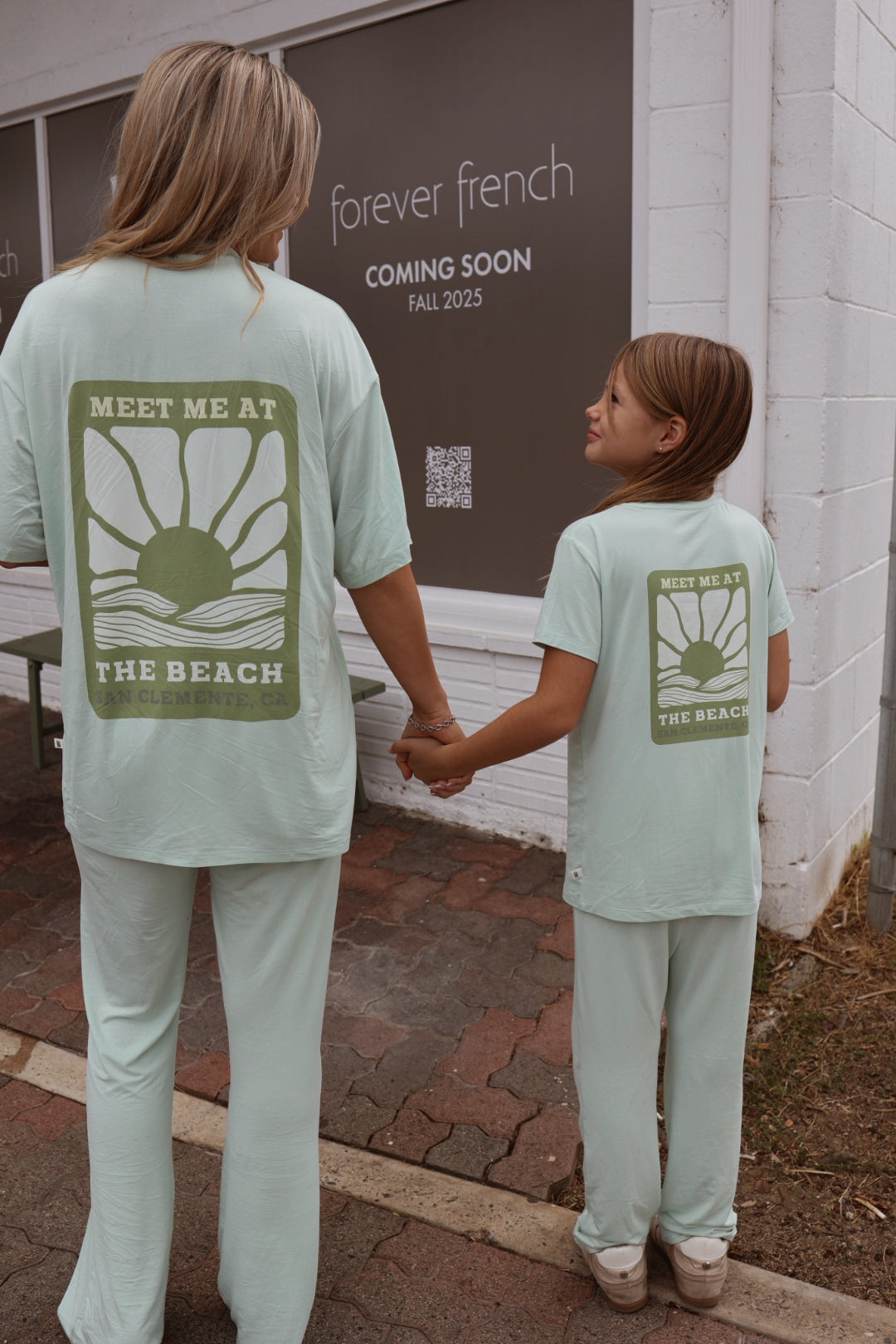 An adult and child wear matching "Meet Me at the Beach" bamboo lounge sets by forever french baby, featuring a sun graphic. A sign behind them announces: “forever french COMING SOON FALL 2025.”.