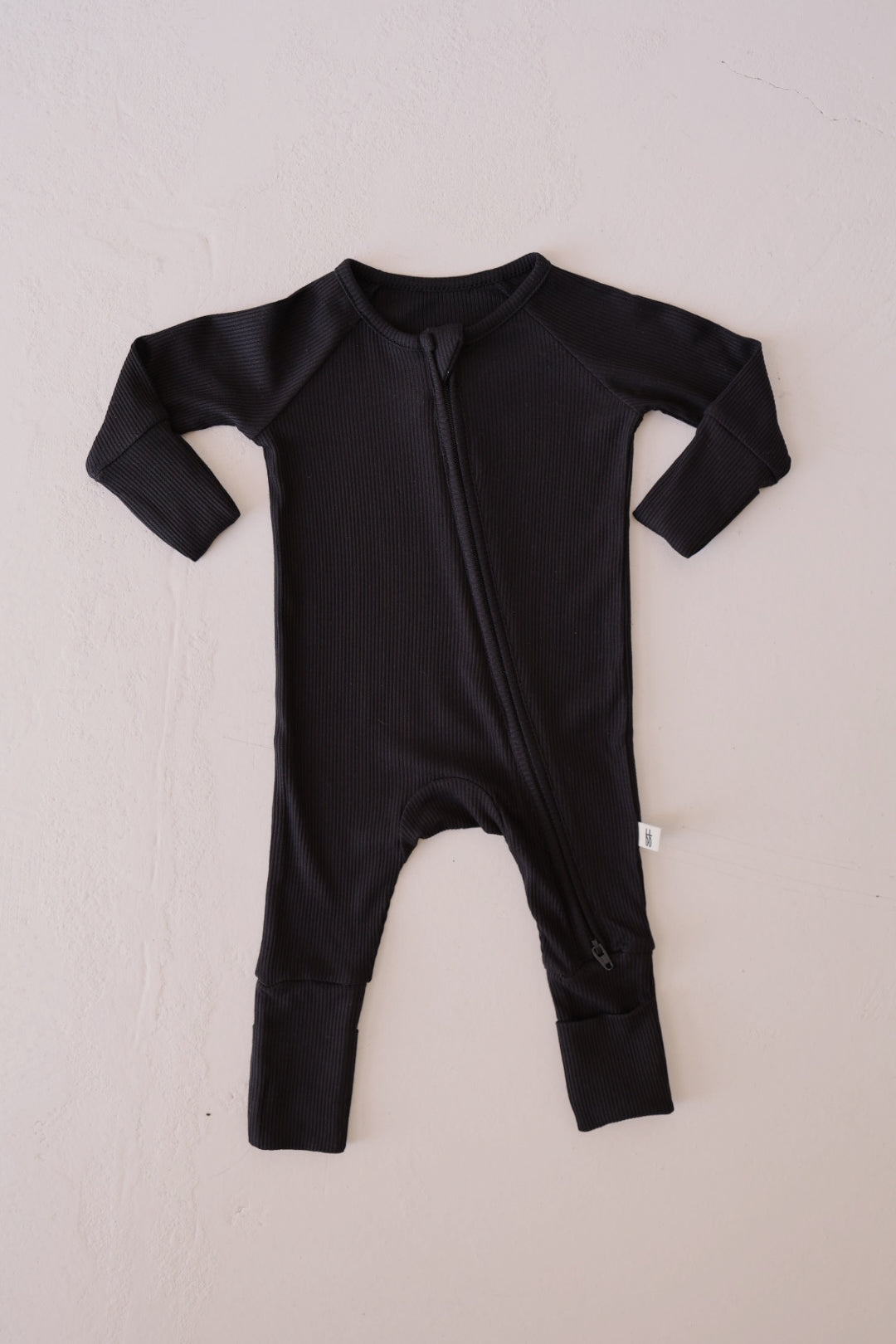 A Black Ribbed Bamboo Zip Pajamas onesie from forever french baby, featuring long sleeves and a diagonal zipper that extends from the left shoulder to the right ankle. Made from hypo-allergenic fabric with a ribbed texture, it includes fold-over cuffs at both the sleeves and legs. The onesie is displayed flat on a light-colored surface.