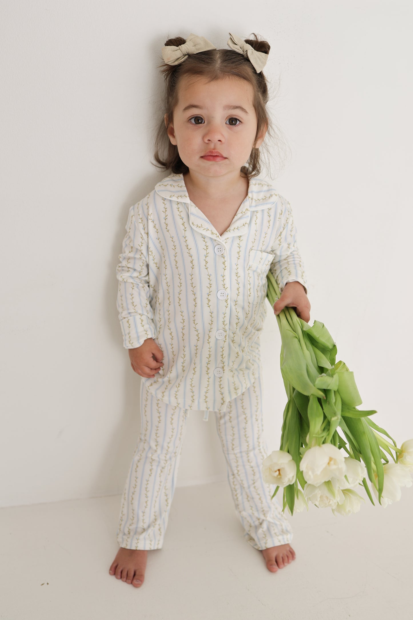 A young girl with bows in her hair, barefoot against a white wall, wears the forever french baby Girl’s Bamboo Button Up Lounge Set | Dainty Vines—hypo-allergenic bamboo clothing ideal for sensitive skin.
