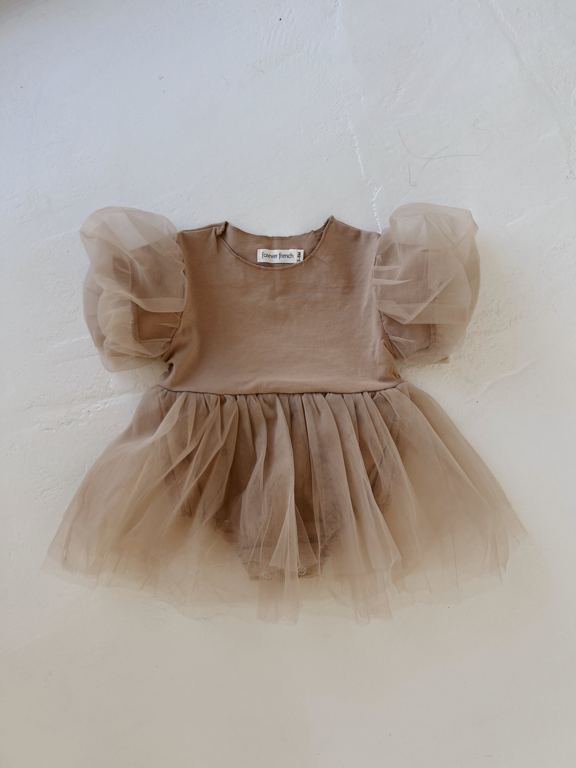 The taupe Puff Sleeve Snap Suit Dress from forever french baby is laid out flat on a light-colored surface. The dress features a plain bodice and puffy sheer sleeves, while the skirt, made of layers of soft, delicate tulle, gives it a whimsical and airy appearance that embodies the charm of a classic puff sleeve design.