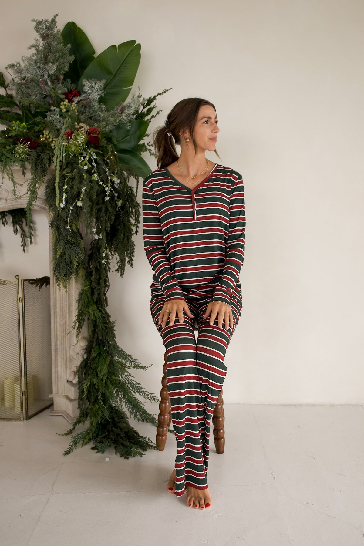 A woman sits wearing a Women’s Long Sleeve Bamboo Pajama | Holiday Stripe set featuring long sleeves, buttons, and horizontal holiday-colored stripes.