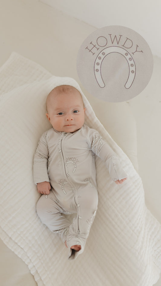 A baby lies on a soft white blanket wearing the "forever french baby" Bamboo Zip Pajamas in Howdy design, featuring light colors, breathable hypo-allergenic bamboo fabric, and adorned with "Howdy" and horseshoe motifs.