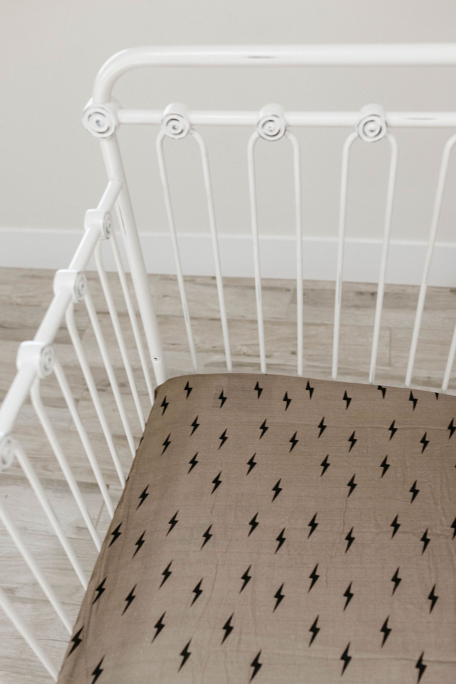 A white vintage-style crib with ornate detailing and a brown mattress, adorned with a Muslin Crib Sheet featuring a pattern of small black lightning bolts from forever french baby, is situated on a light wood floor against a beige wall.