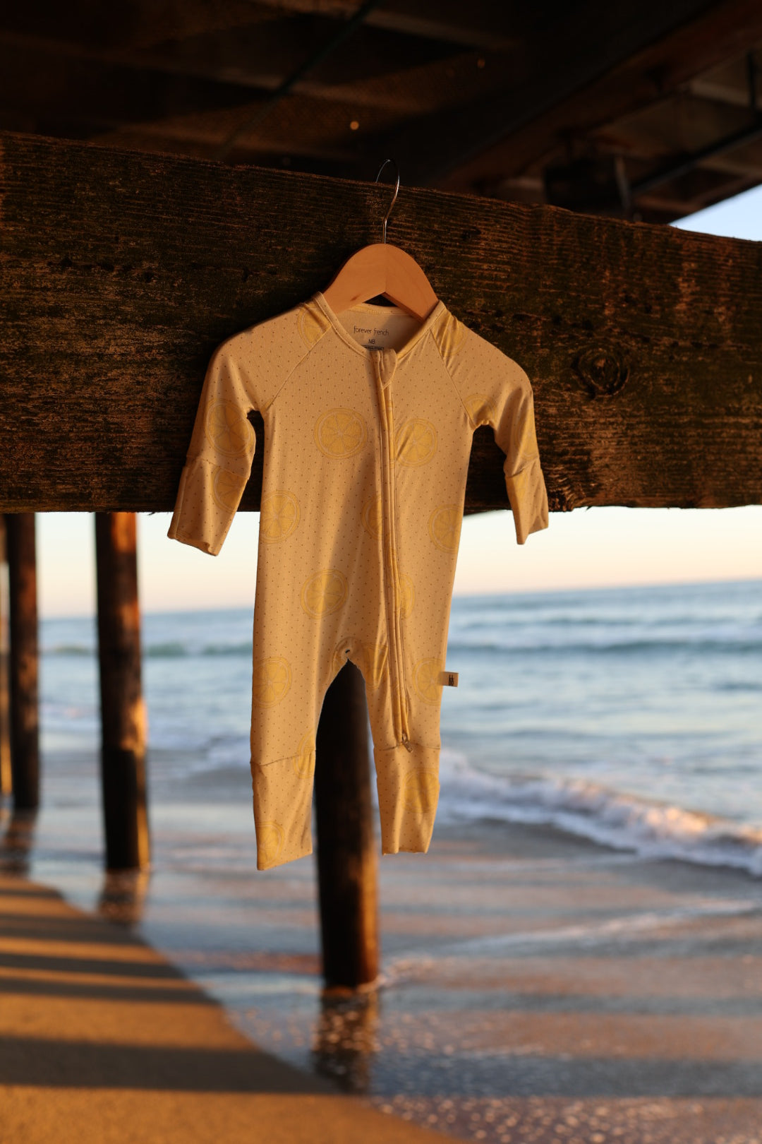 A yellow Bamboo Zip Pajama (Lemon Drop) from forever french baby hangs from a pier beam at sunset on the beach—gentle waves in the background highlight this perfect, breathable, hypoallergenic sleepwear for your little one.