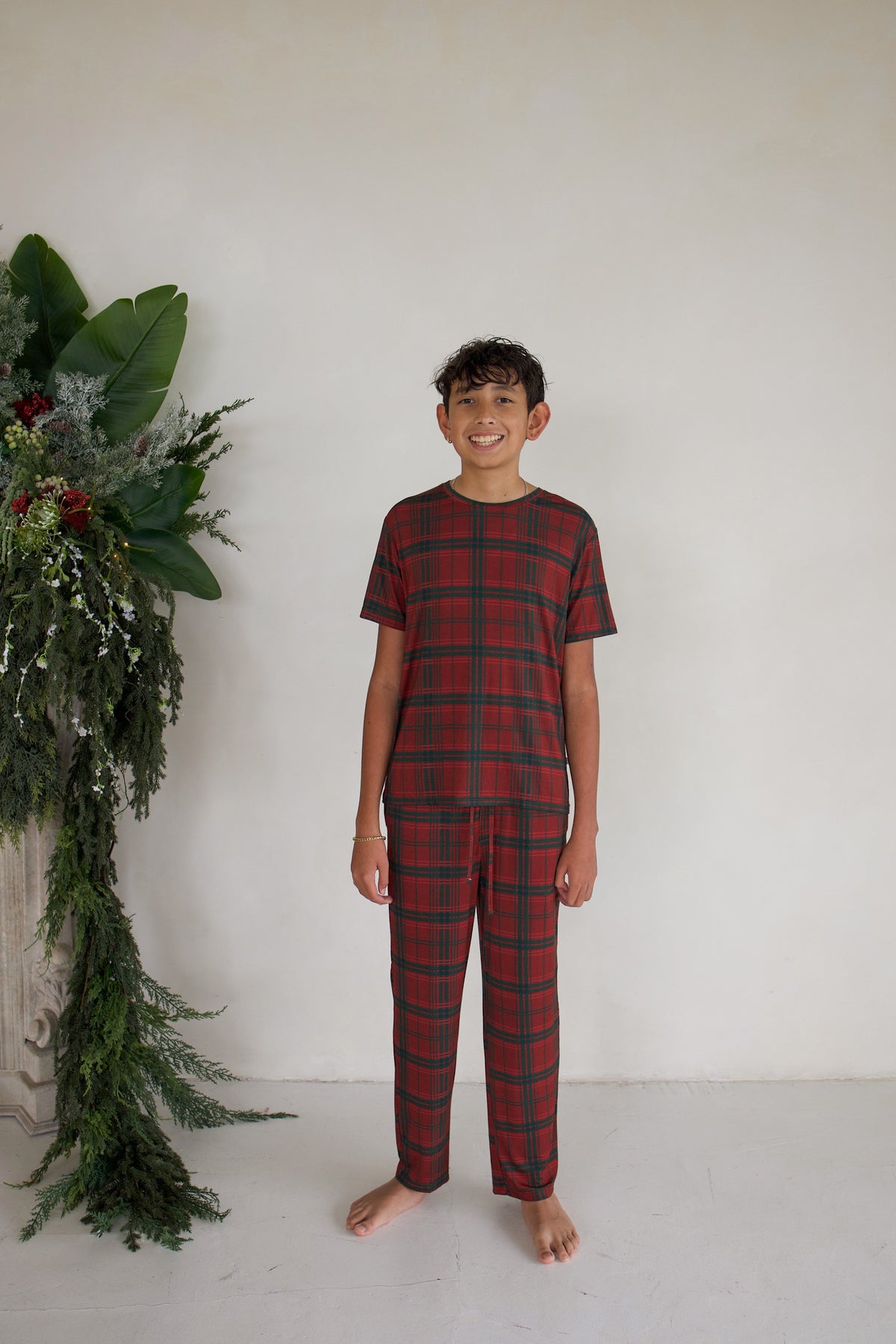A barefoot boy wears short-sleeved red and green Christmas Plaid Youth Straight Leg Bamboo Pajamas by forever french baby.
