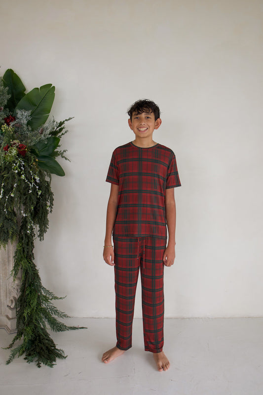 A barefoot boy wears short-sleeved red and green Christmas Plaid Youth Straight Leg Bamboo Pajamas by forever french baby.