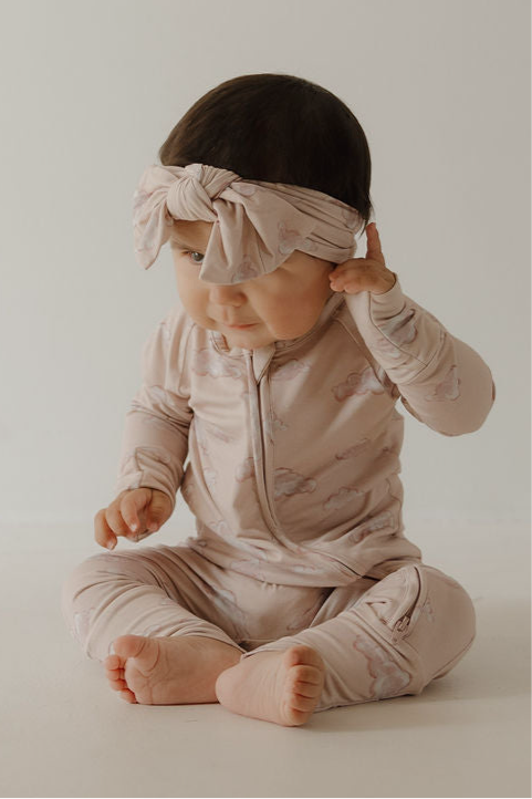A baby sits on the floor wearing Kendy 3’s Bamboo Zip Pajamas in Ruby with a matching large bow headband, looking down and touching one ear.