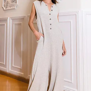 A person stands in a hallway with white wainscoting, wearing the Bucketlist Wide Leg Button-Up Loose Fit Solid Sweater Jumpsuit. The light-colored garment boasts a subtle texture and wide legs. With one hand casually tucked into their pocket, the individual exudes a relaxed vibe.