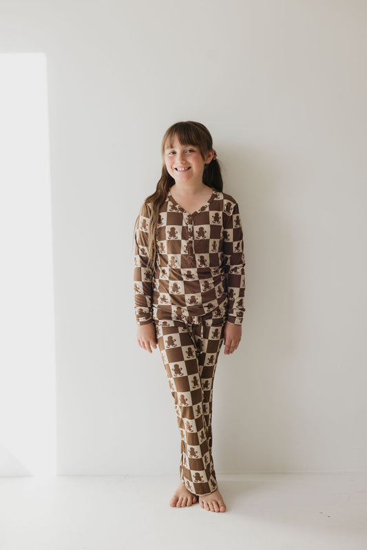 A young girl stands against a white wall, smiling in her "Teen Bamboo Pajamas | Gingerbread Checkerboard" from forever french baby. The minimal background and brightly lit floor highlight her hypo-allergenic clothing, perfect for sensitive skin.