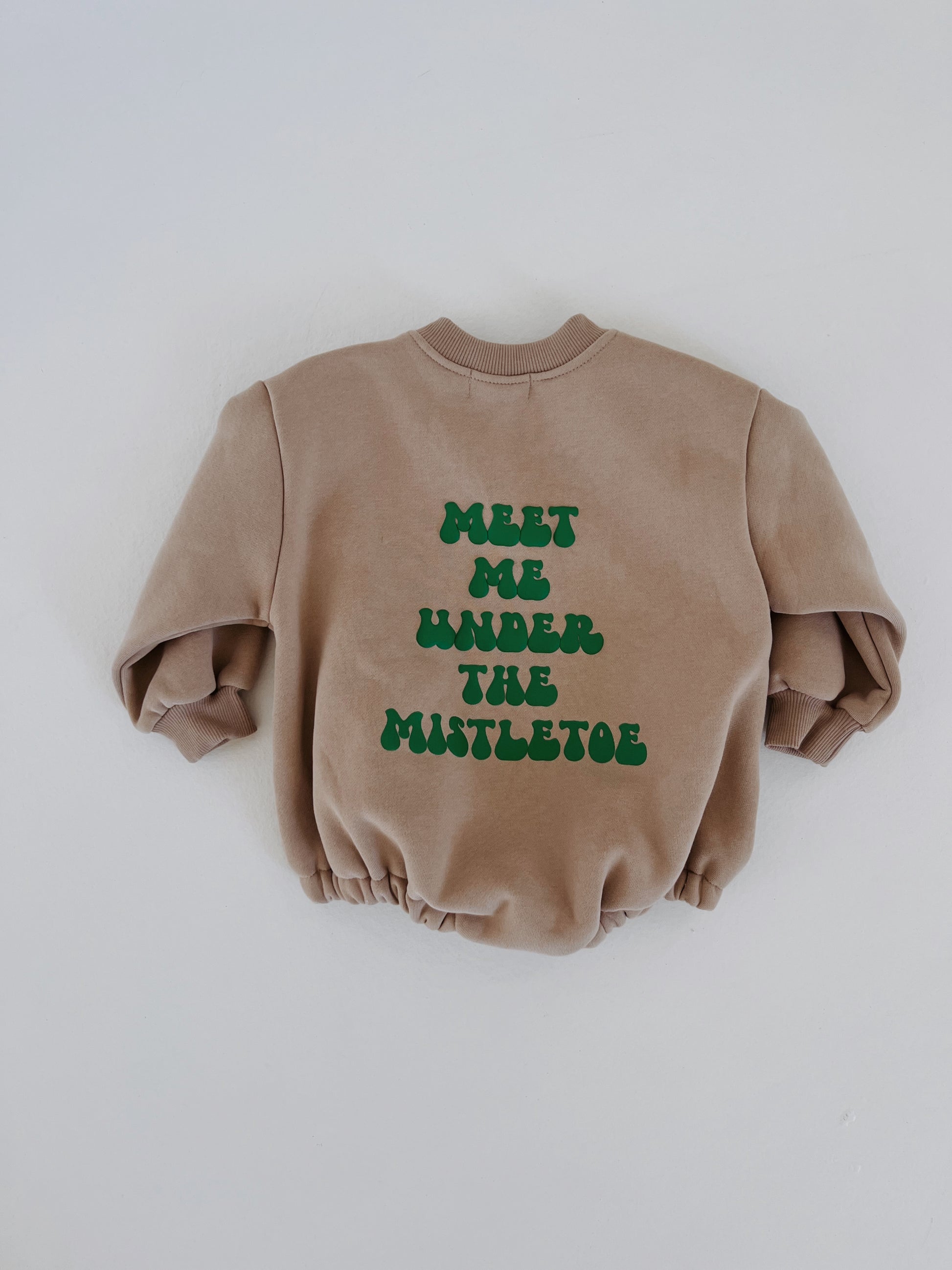 A light brown long sleeve sweatshirt romper from forever french baby is showcased under soft lighting, exuding holiday charm. It features green "MEET ME UNDER THE MISTLETOE" text in a playful font on the back, perfect for cozy moments and fans of the brand.