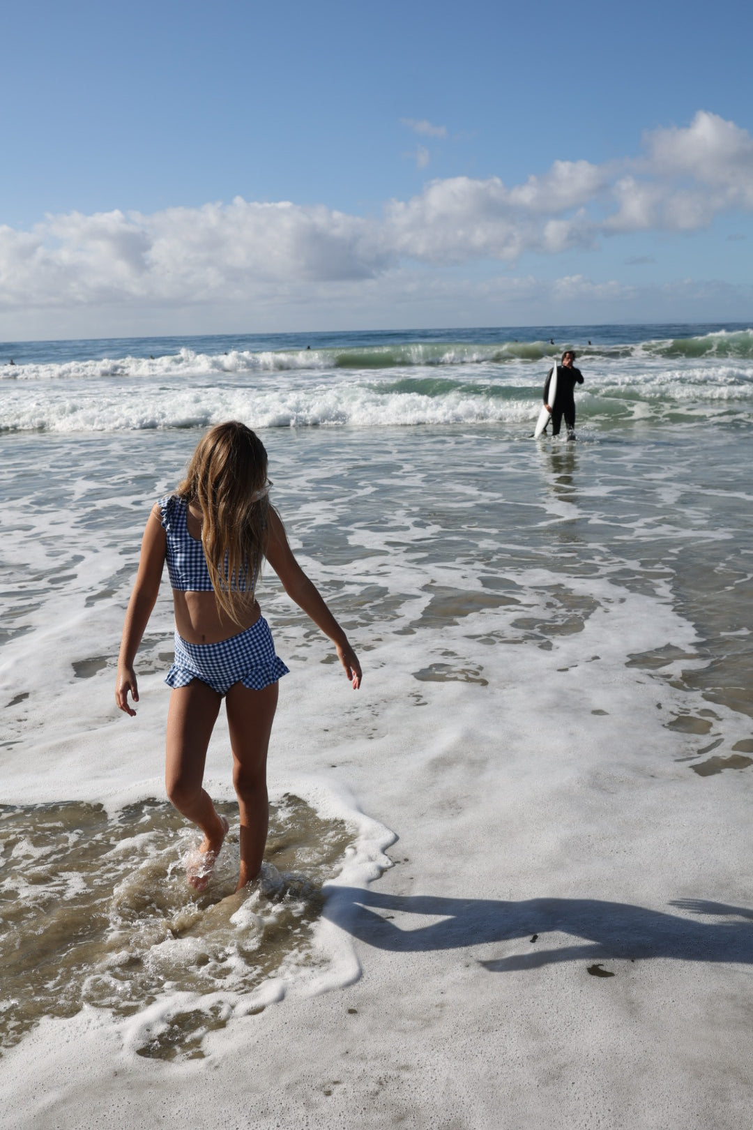 A girl wearing the forever french baby Girls Two Piece Swimsuit in Blue Gingham walks by the ocean, while a surfer stands in the waves under a blue, cloud-dotted sky.