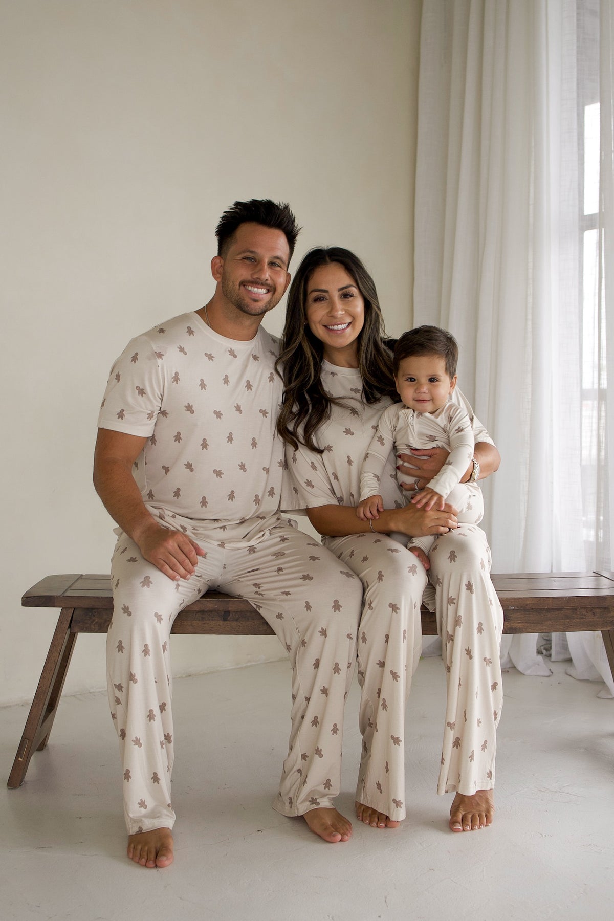 A family of three sits on a wooden bench wearing matching beige bamboo Men’s Short Sleeve Pajamas | Gingersnap featuring a small gingerbread man print.
