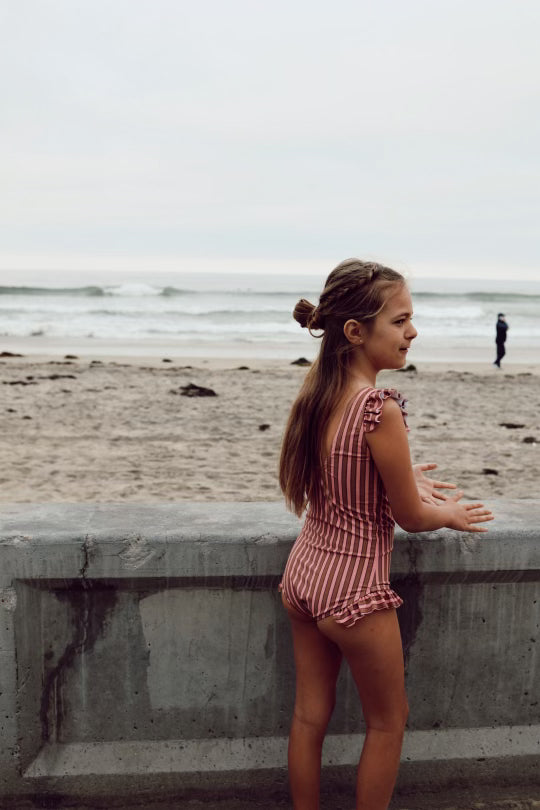 A young girl with long hair stands at a concrete barrier, facing the beach and ocean. Clad in a Boardwalk One Piece Girls Swim Suit from forever french baby, which features earth tone stripes and UPF 50+ protection, she gazes towards the water on a cloudy day. Another person can be seen in the distance near the shoreline.