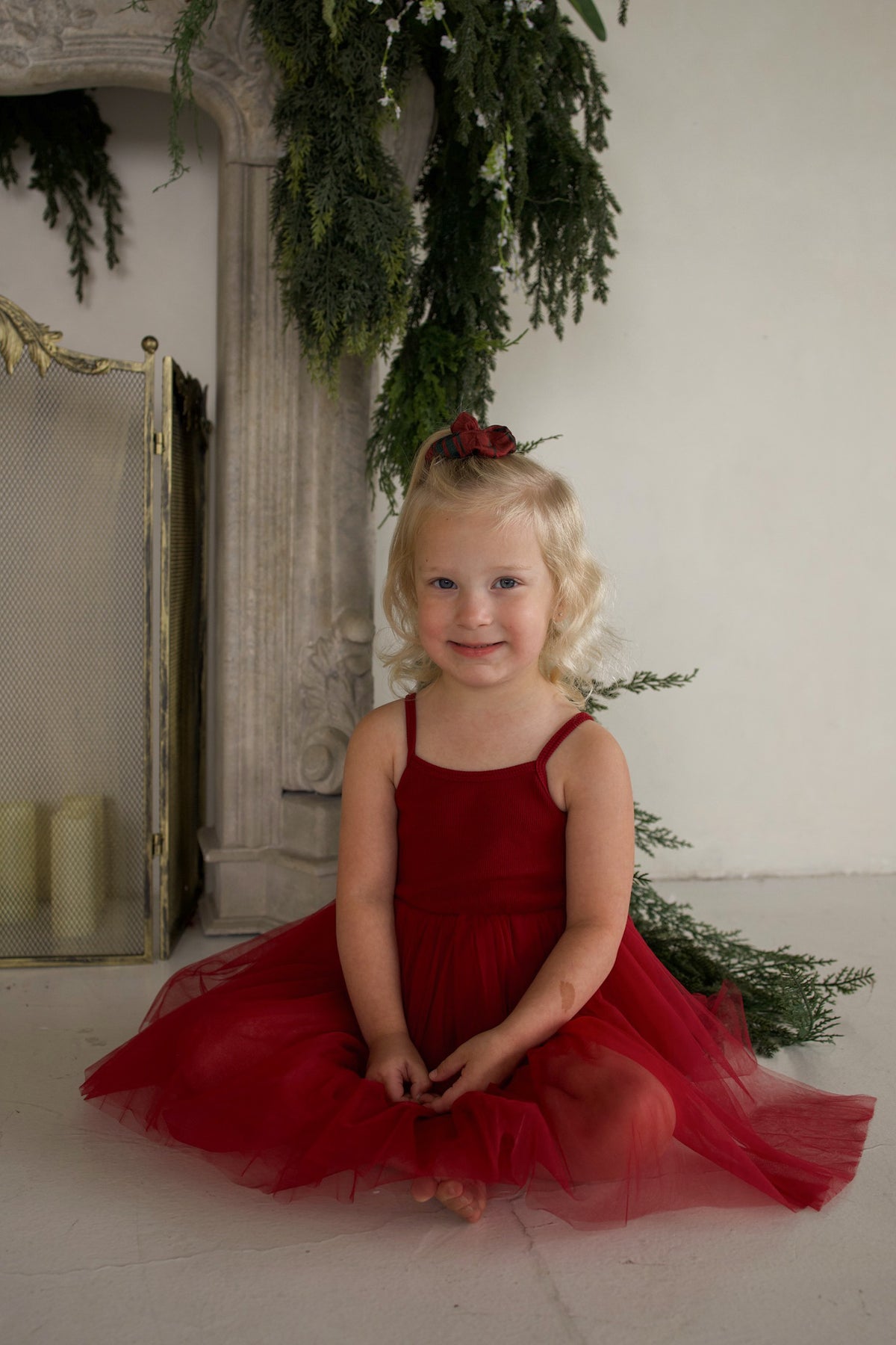 A young girl sits on the floor wearing the forever french baby ff Tutu Dress | Cranberry, featuring a sleeveless design and a layered tulle skirt.