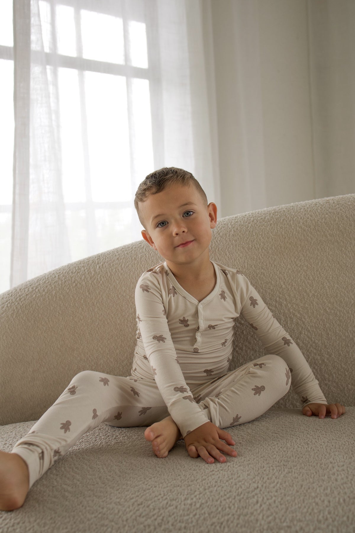 A young boy sits on a couch wearing Bamboo Two Piece Pajamas | Gingersnap, featuring a long-sleeved, breathable gingerbread man print design.