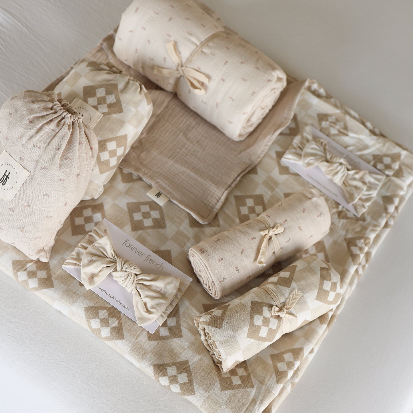 Forever french baby's Muslin Swaddle | Cottage Quilt in beige and cream patterns, neatly folded on a matching sheet—some tied with ribbon, others in drawstring bags—perfect for cozy, stylish nursery decor.