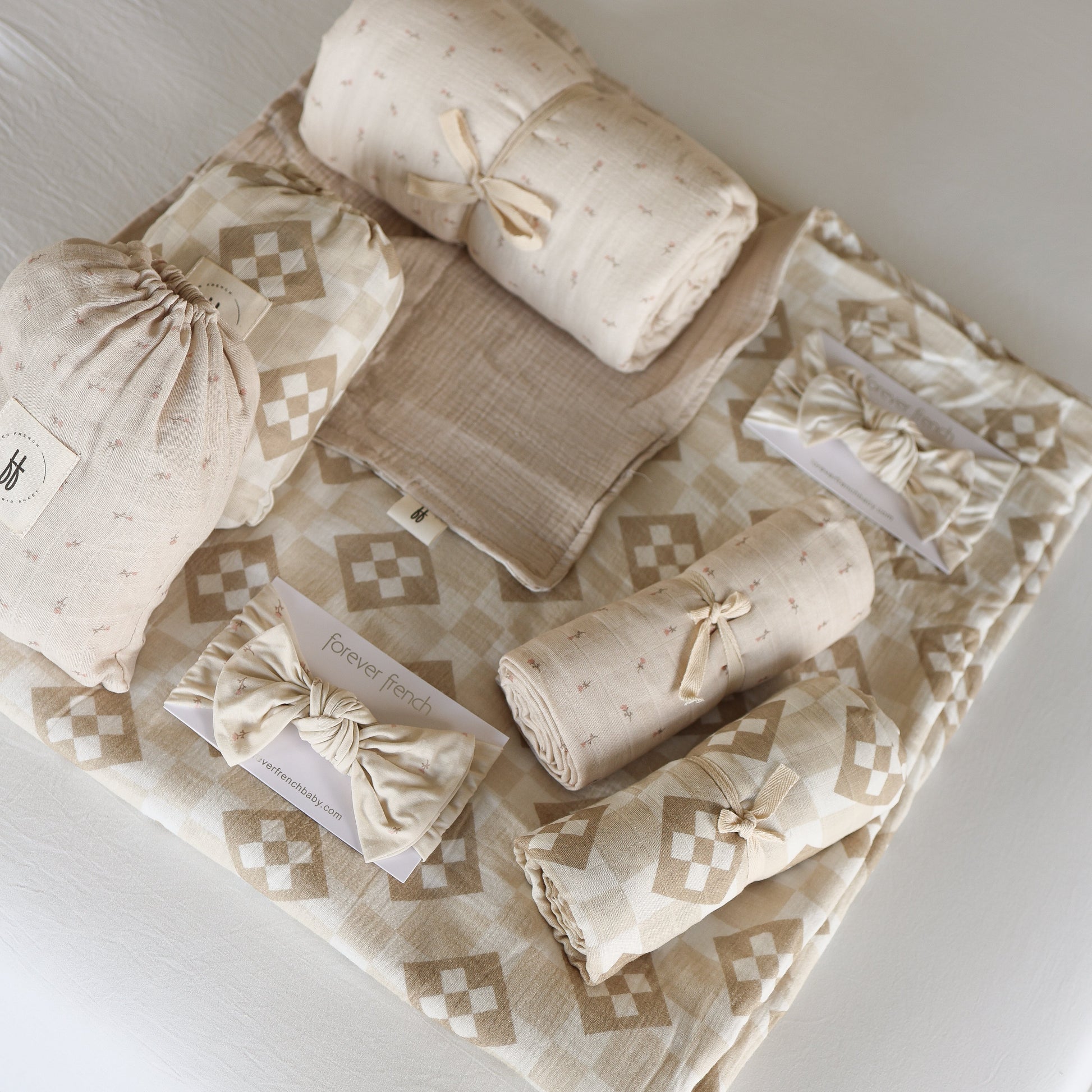 Forever french baby's Muslin Swaddle | Cottage Quilt in beige and cream patterns, neatly folded on a matching sheet—some tied with ribbon, others in drawstring bags—perfect for cozy, stylish nursery decor.