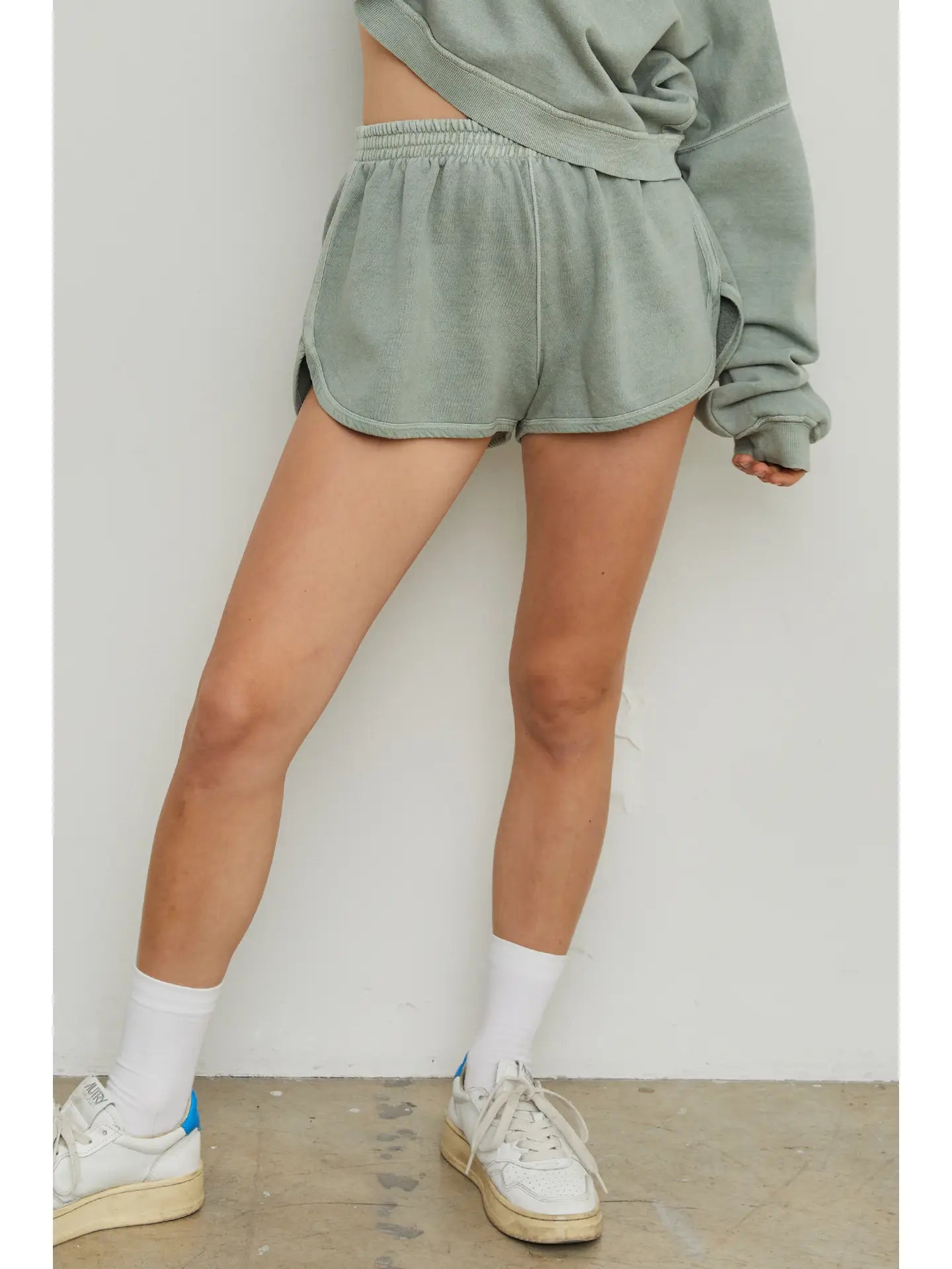 A person wearing a sage green sweatshirt and matching Organic Cotton Shorts | Sage Green from forever french baby, pulling up one sleeve, with white socks and off-white sneakers, is standing with one leg slightly bent against a light-colored wall. The floor appears to be concrete.