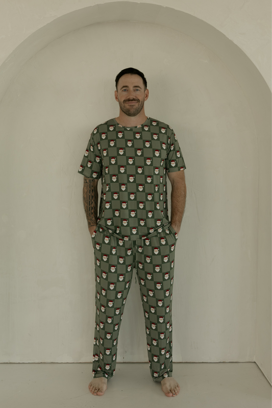 A man with short dark hair and a beard stands barefoot in a neutral space, wearing forever french baby’s Men's Short Sleeve Bamboo Pajamas in the Santa Checkerboard pattern, hands in his pockets and smiling at the camera.