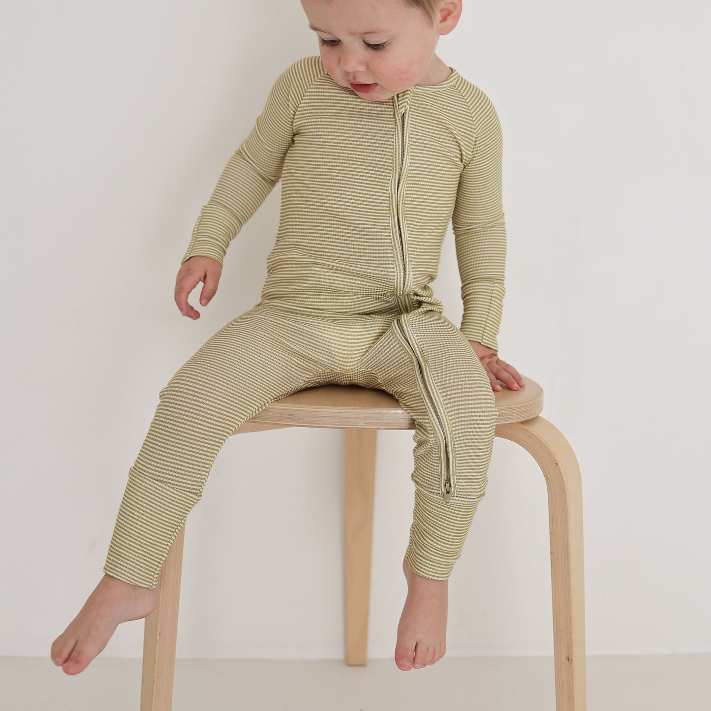 A toddler in forever french baby’s Bamboo Zip Pajamas | Willow Stripe Ribbed sits on a light wooden stool against a plain white background, looking down and touching the seat.