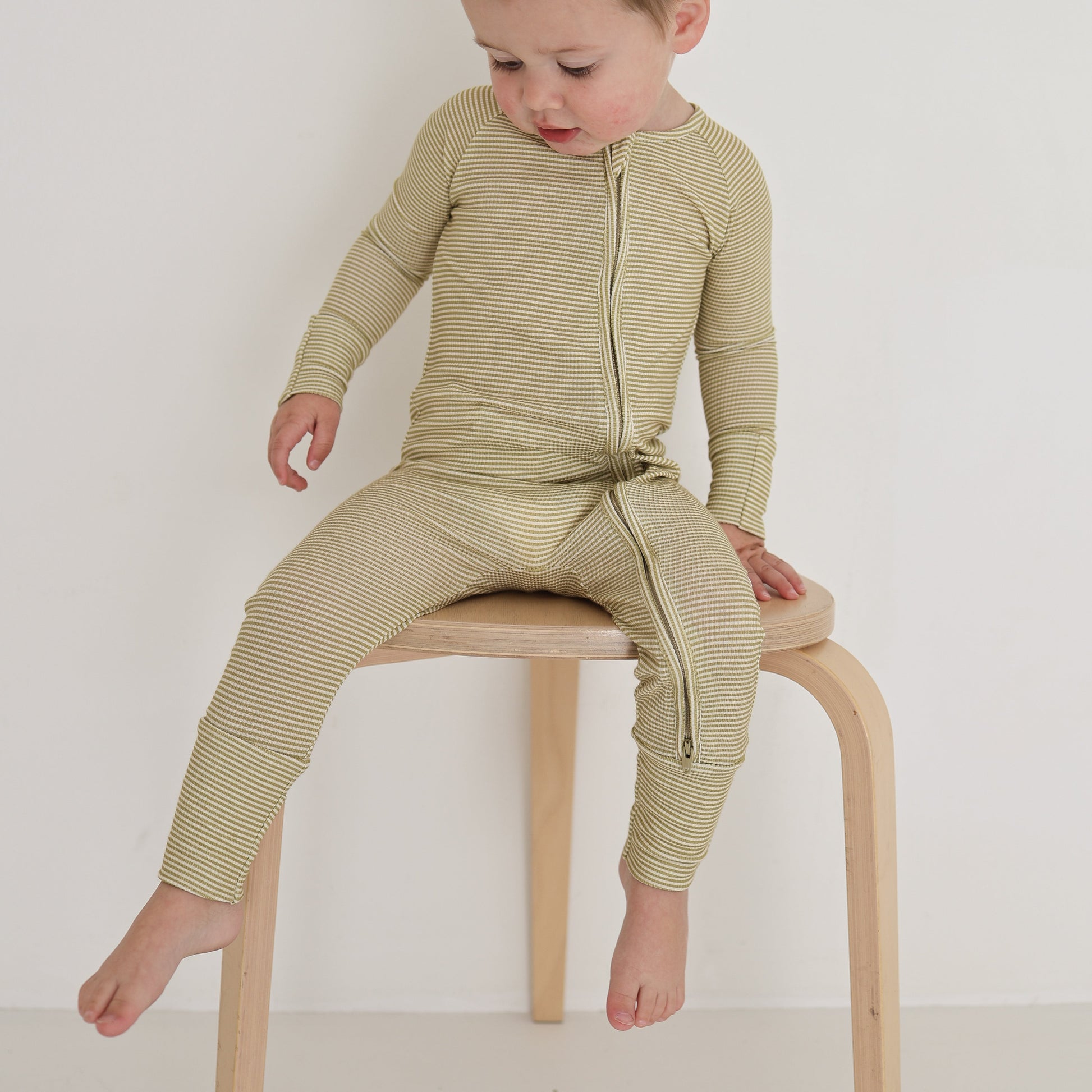 A toddler in forever french baby’s Bamboo Zip Pajamas | Willow Stripe Ribbed sits on a light wooden stool against a plain white background, looking down and touching the seat.