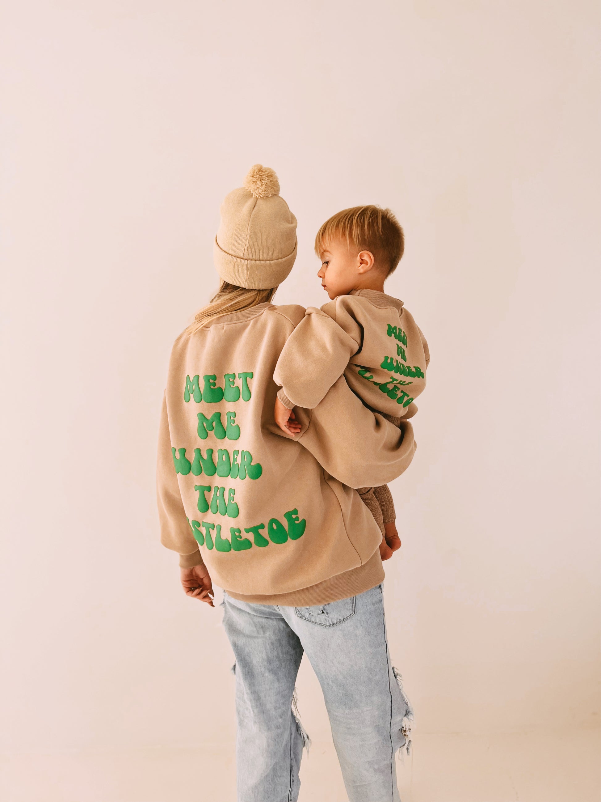 A woman wearing a matching beige beanie and a festive "Meet Me Under The Mistletoe" sweatshirt by forever french baby holds a young child dressed in similar holiday apparel. Both sweatshirts feature green text on the back, hers reading "MEET ME UNDER THE MISTLETOE" and the child's "MINI UNDER THE MISTLETOE.