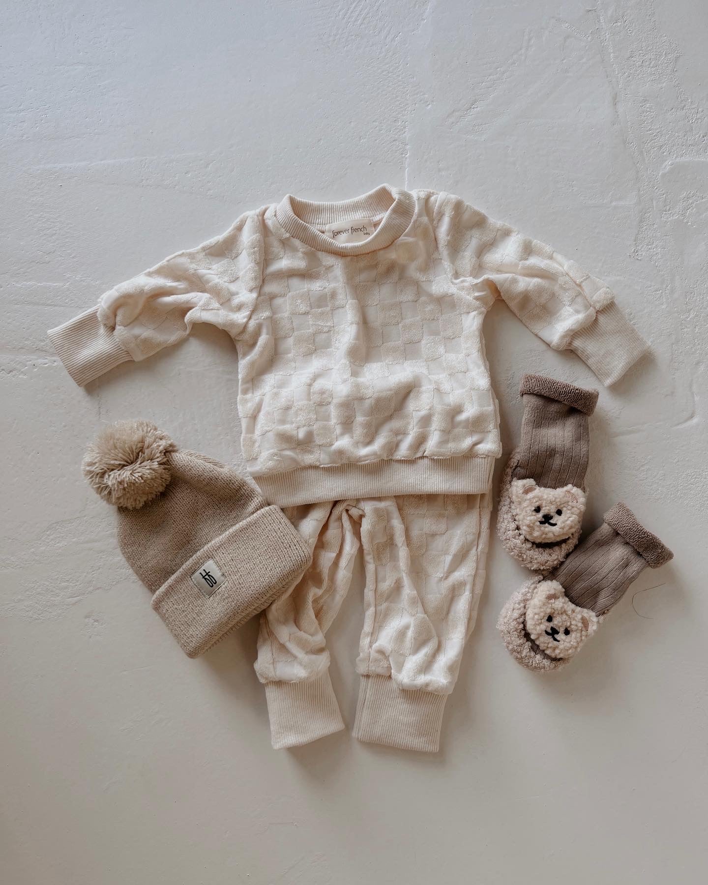 A set of baby clothing is laid out on a light surface, featuring a cream-colored knit sweater and pants with a checkerboard pattern, a beige knit hat with a pom-pom, and an adorable pair of Teddy Bear Slipper Socks by forever french baby in beige and brown with bear faces on the front.