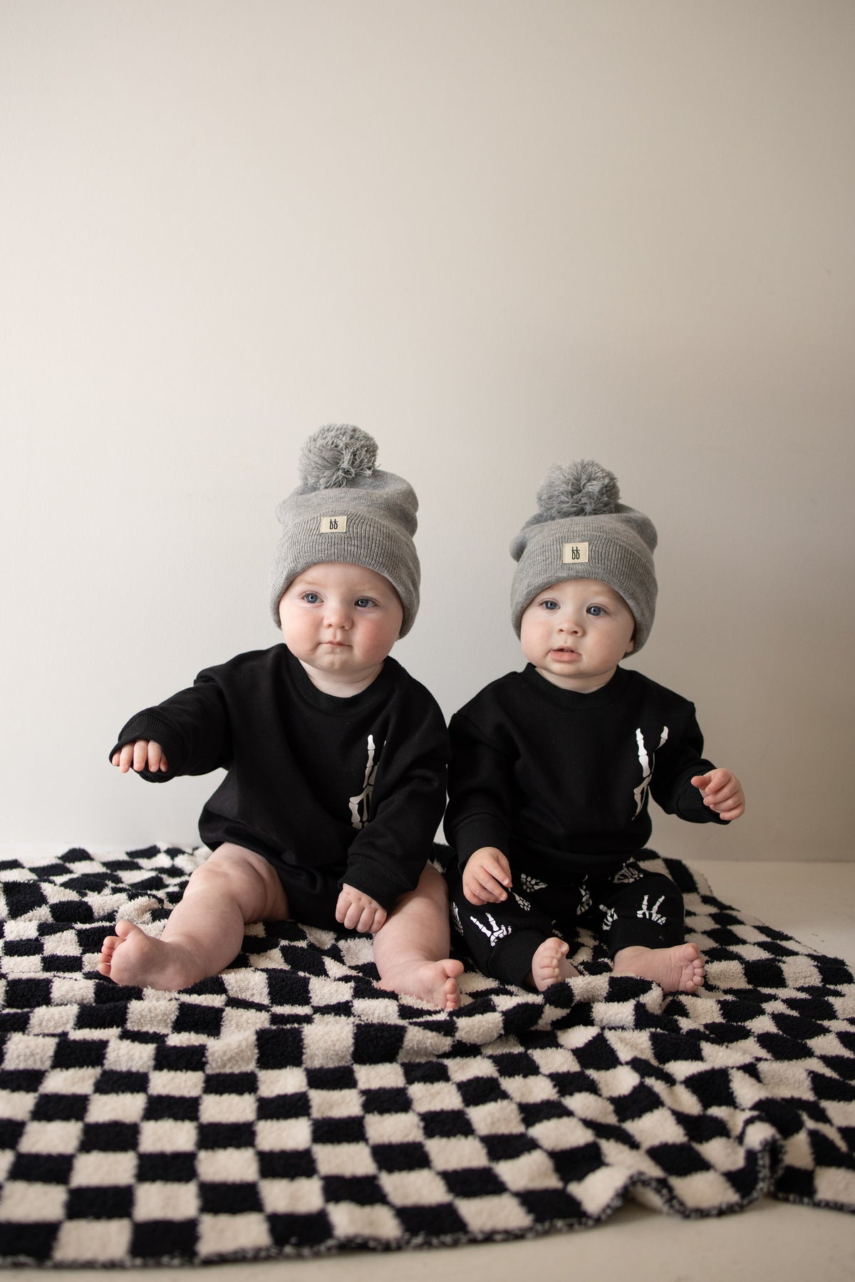 Two babies in matching gray pom-pom beanies and black Sweatshirt Romper | Midnight Skelly outfits from forever french baby sit on a black-and-white checkered blanket, looking toward the camera against a plain light background.