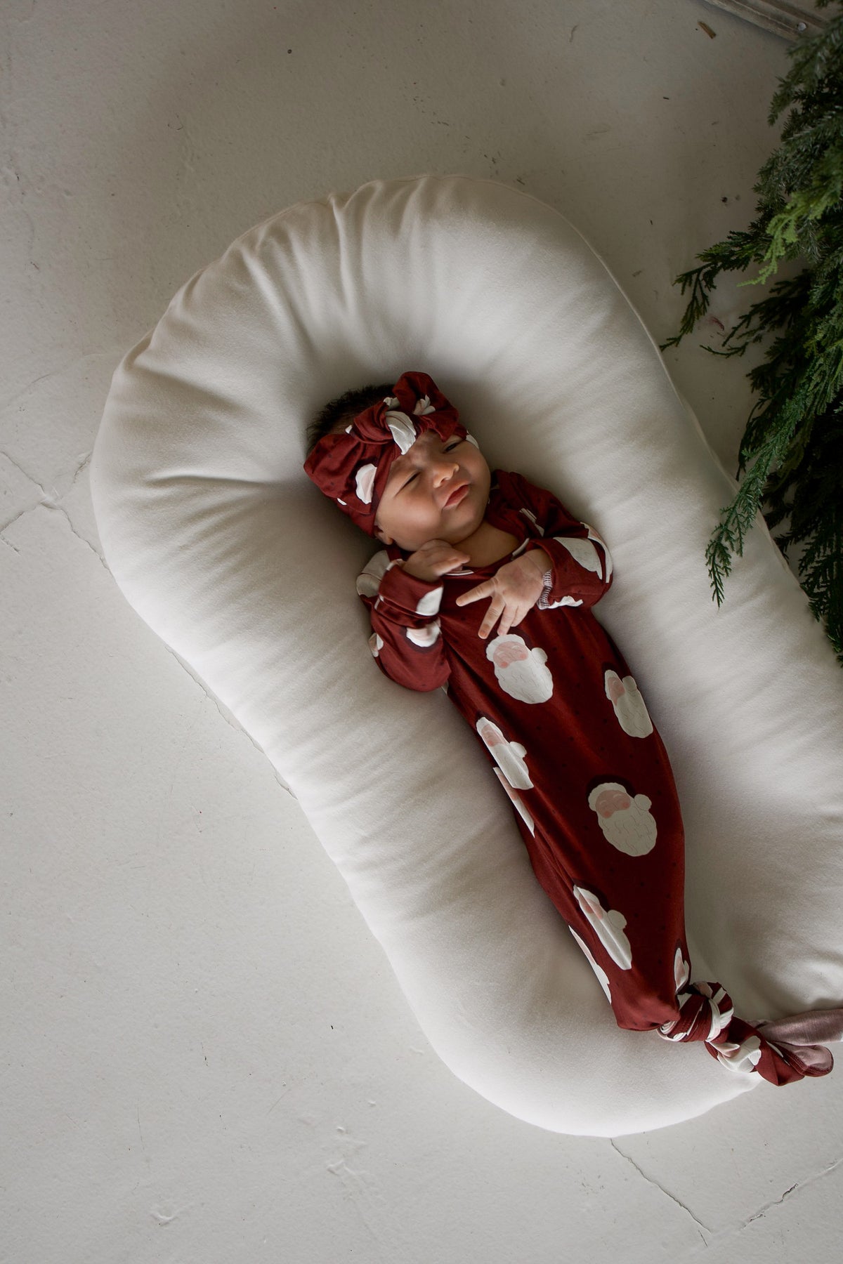 A newborn wearing a Bamboo Knotted Gown | Vintage Santa in dark red with a matching bow headband, both patterned with Santa Claus faces, resting in a white baby lounger.