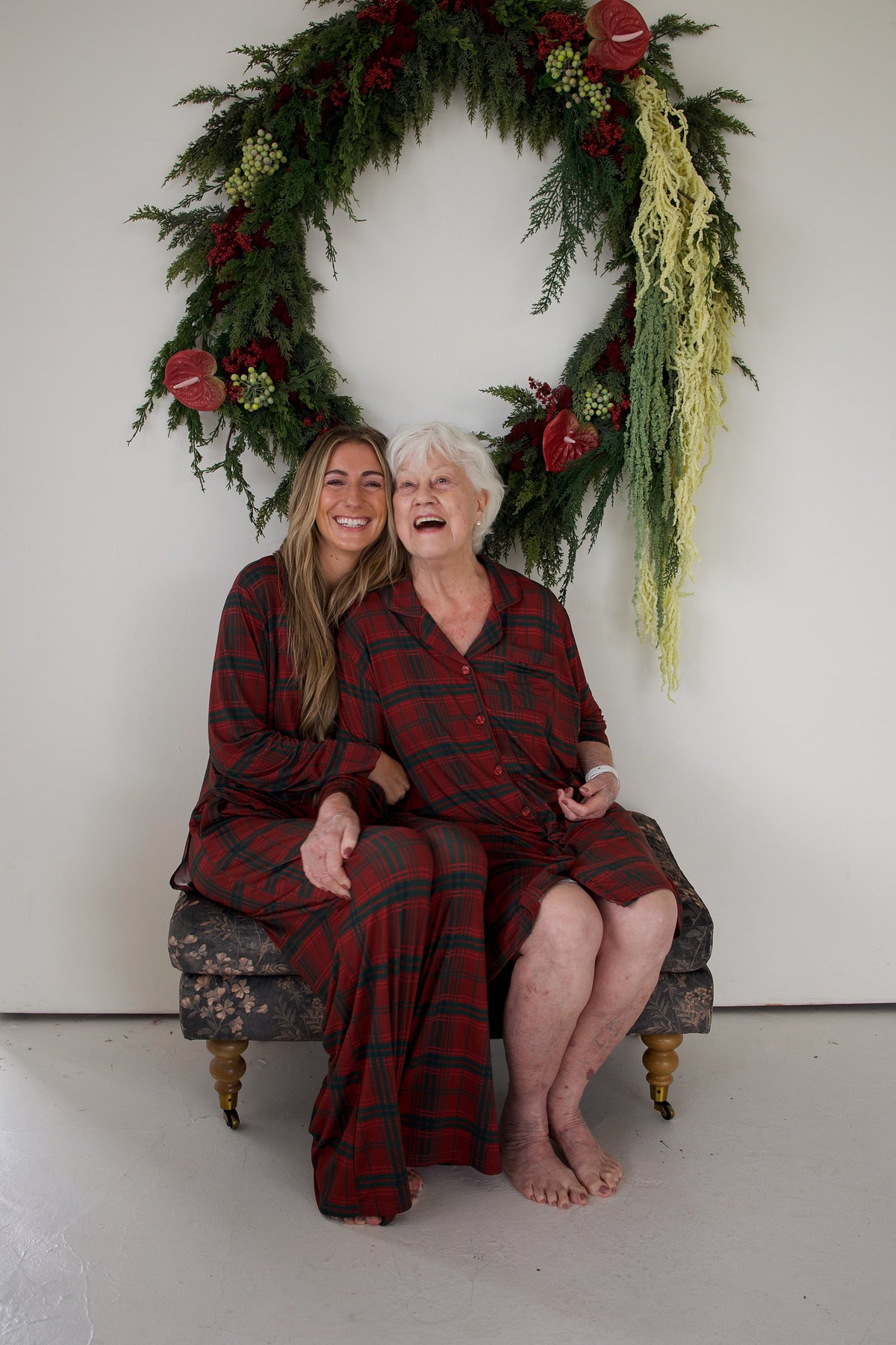 Two women laugh while wearing matching Women’s Bamboo Sleeping Dress | Christmas Plaid, sitting on an upholstered bench in front of a holiday wreath.