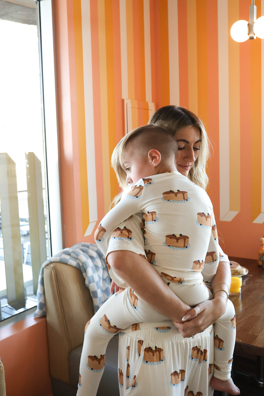 A woman hugs a young child, both wearing matching forever french baby Bamboo Two Piece Pajamas | Sunday Stack with a dog print, in a cozy room with orange and white striped walls and natural light from a window.