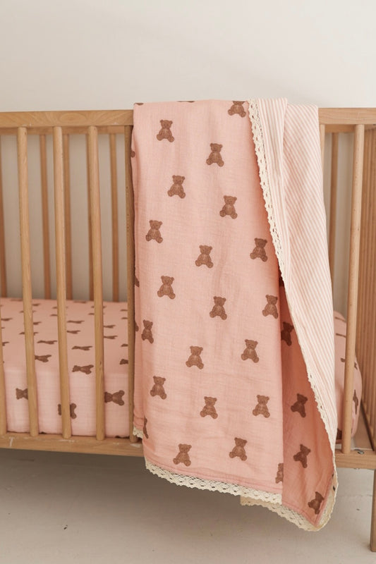 A wooden crib with nursery bedding is adorned with a forever french baby Muslin Quilt | Pink Lottie & Sadie Stripe draped over the side. The matching pink sheet on the mattress adds to the cozy and simple atmosphere.